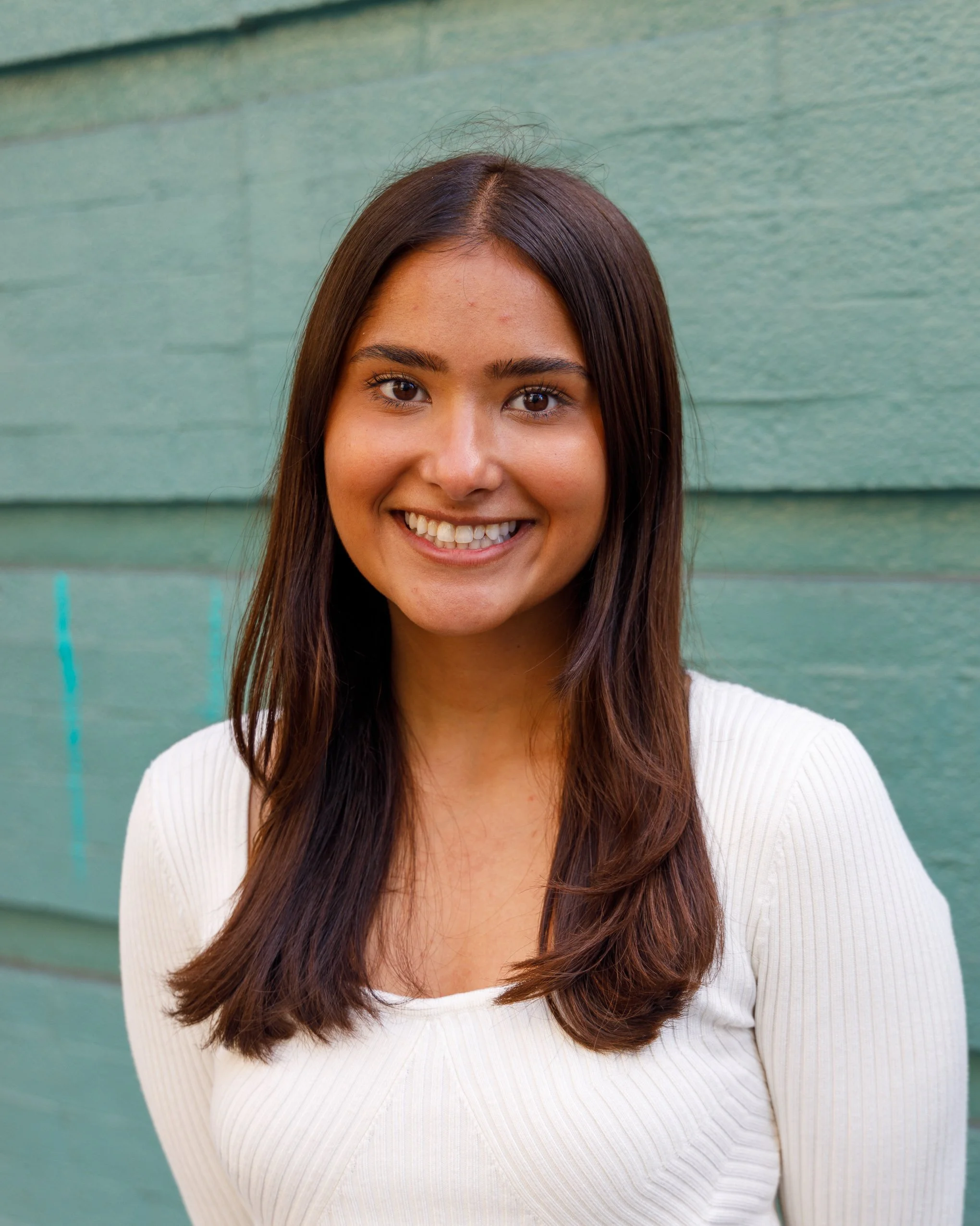 Young woman with long brown hair smiling in front of green wooden wall.