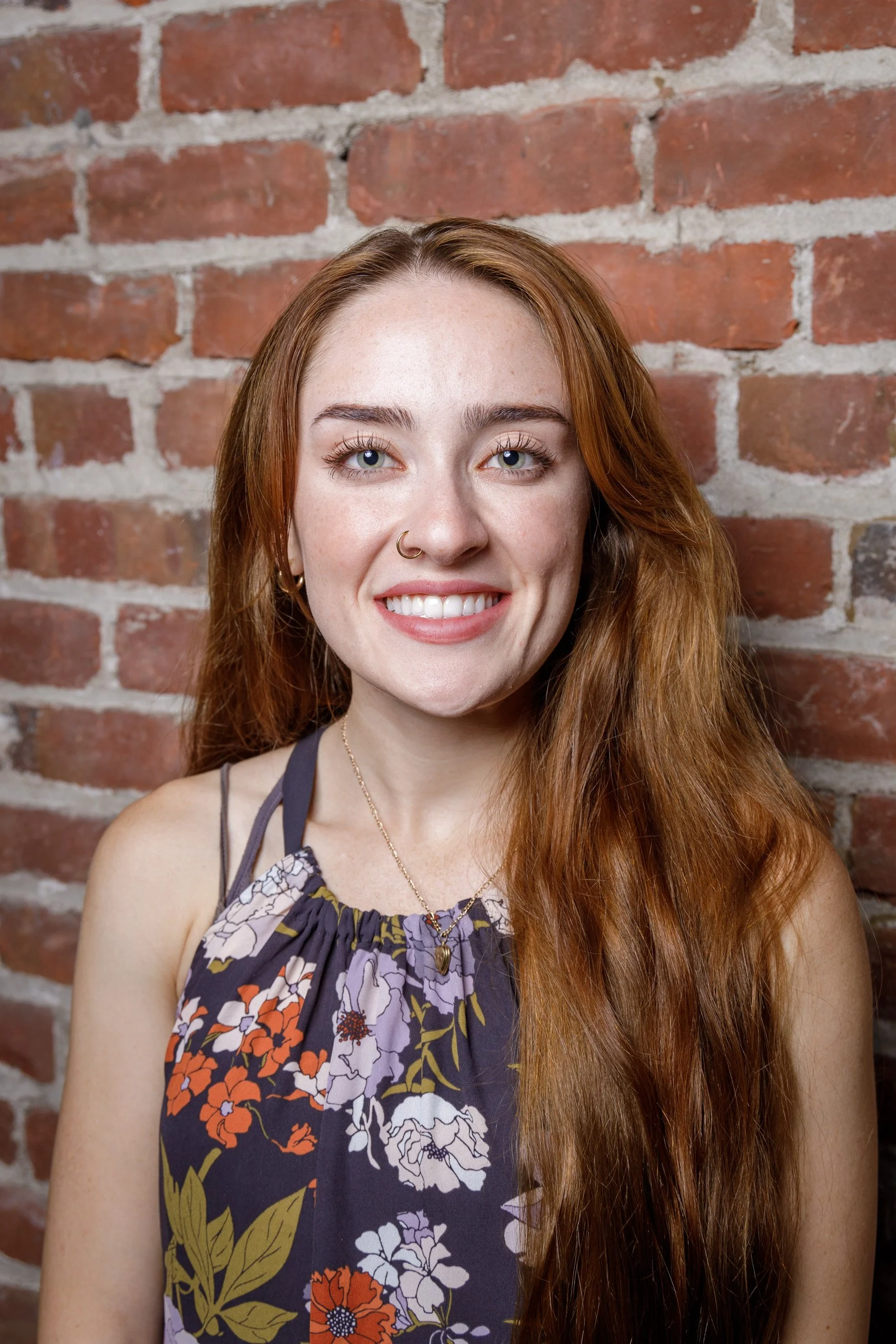 A young woman with long red hair, blue eyes, and a nose piercing smiling at the camera against a brick wall background.