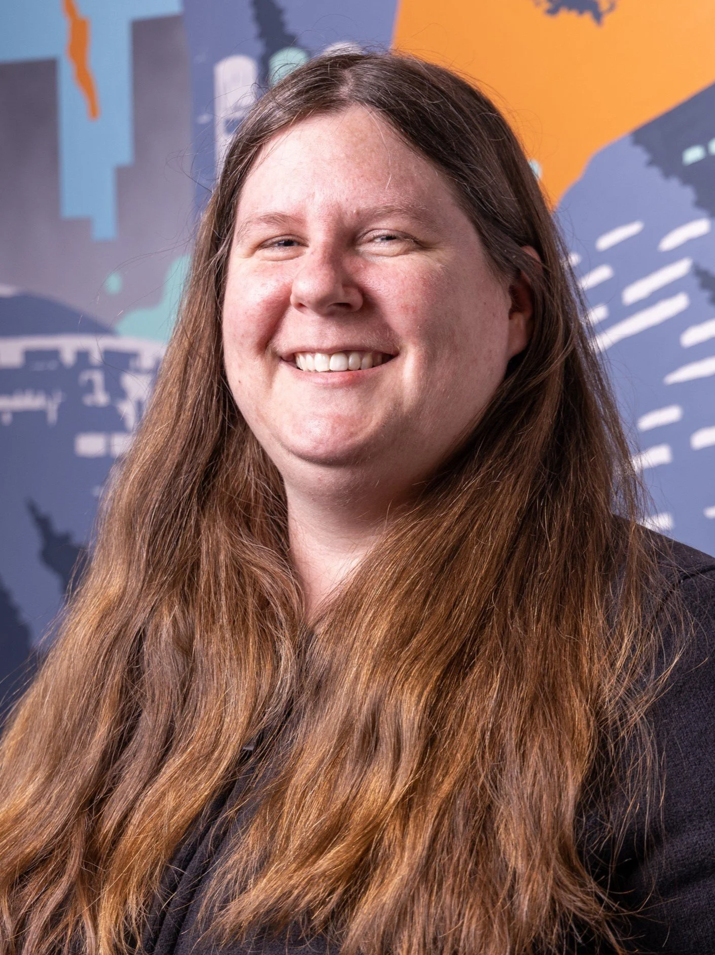 Portrait of a woman with long brown hair, smiling, standing in front of a colorful abstract mural.