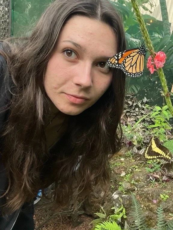 Woman with long curly hair and light skin near butterflies and plants in a garden or zoo.