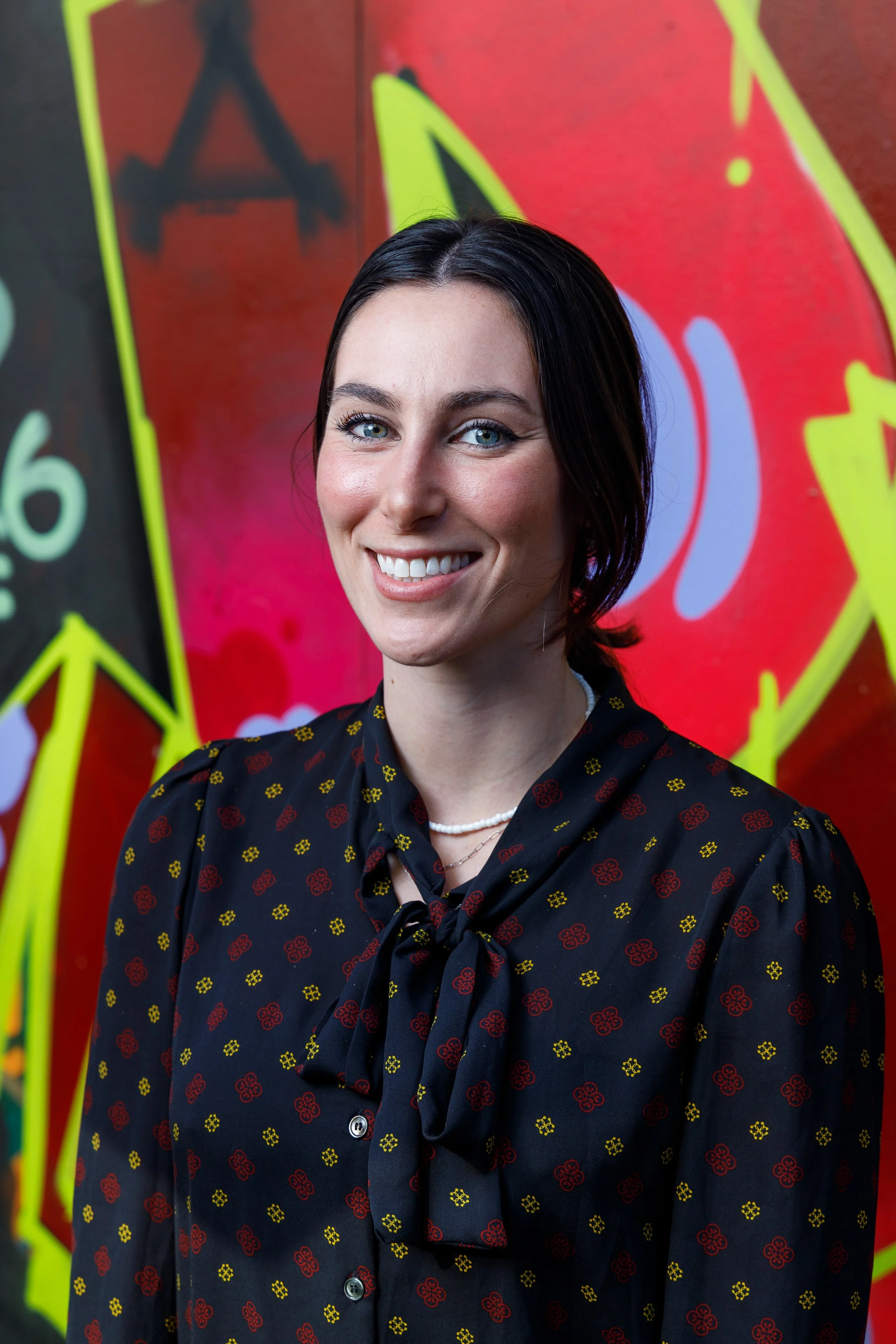 A woman smiling in front of a colorful graffiti wall with spray-painted designs including star and arrow shapes.