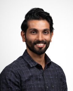 A man with dark hair, a beard, wearing a dark patterned button-up shirt, smiling against a plain light gray background.
