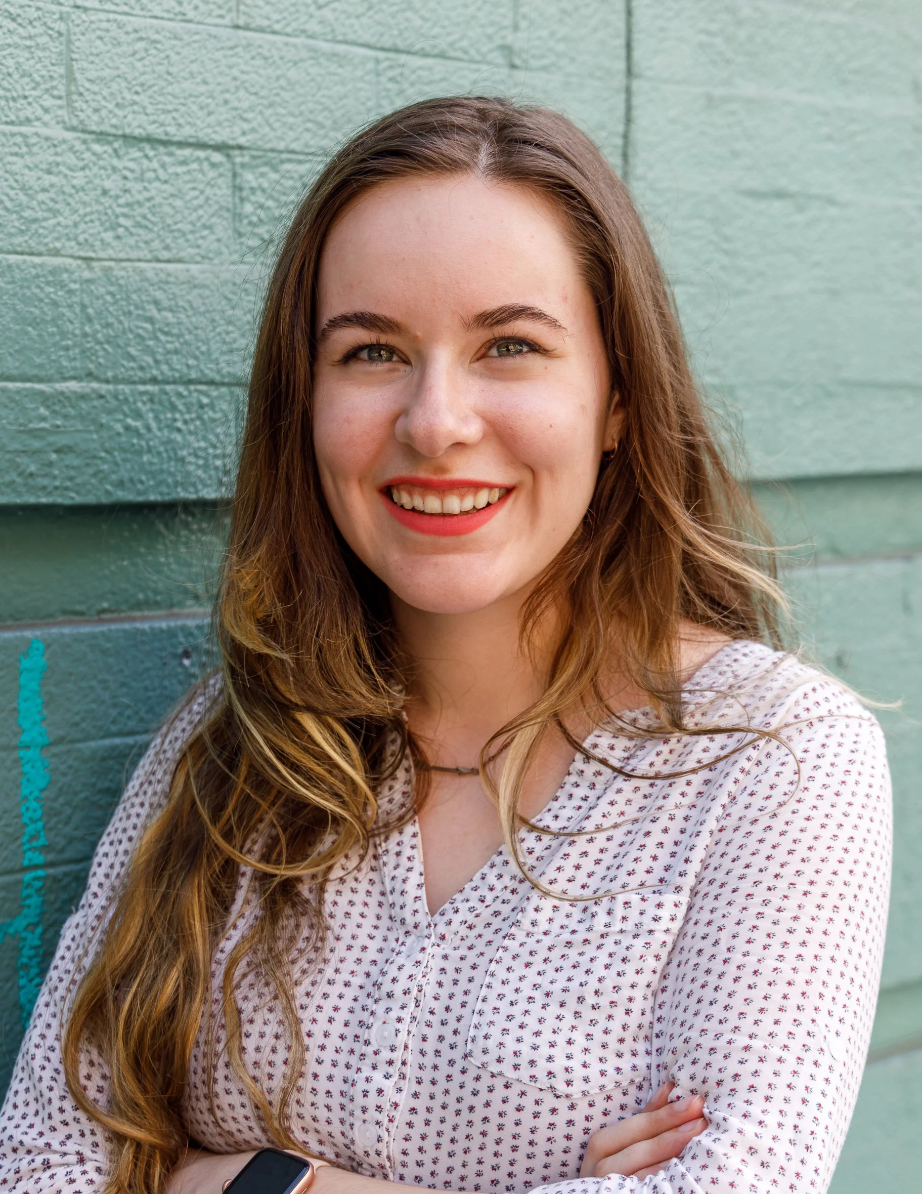 A young woman with long wavy light brown hair, smiling, wearing a white blouse with small red pattern, standing against a green brick wall.