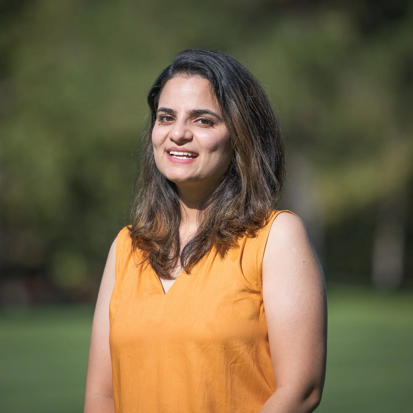 A woman with dark brown, shoulder-length hair wearing a sleeveless orange top standing outdoors. She is smiling and looking at the camera with a blurred green background.