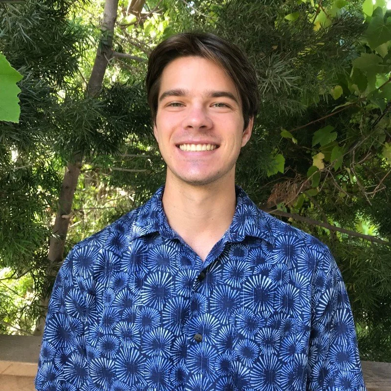 A young man with dark hair and fair skin, smiling, wearing a blue patterned shirt, standing outdoors with green foliage in the background.