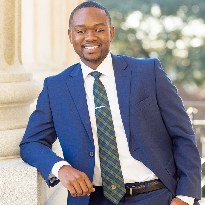 Young man in a blue suit smiling outdoors, leaning against a stone structure with trees in the background.