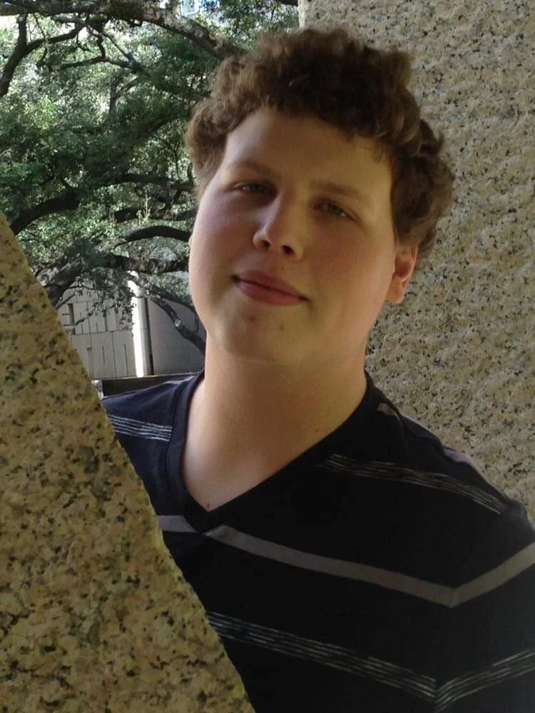 A young man with curly brown hair making a funny face, sticking out his tongue, outdoors surrounded by trees and rocks.