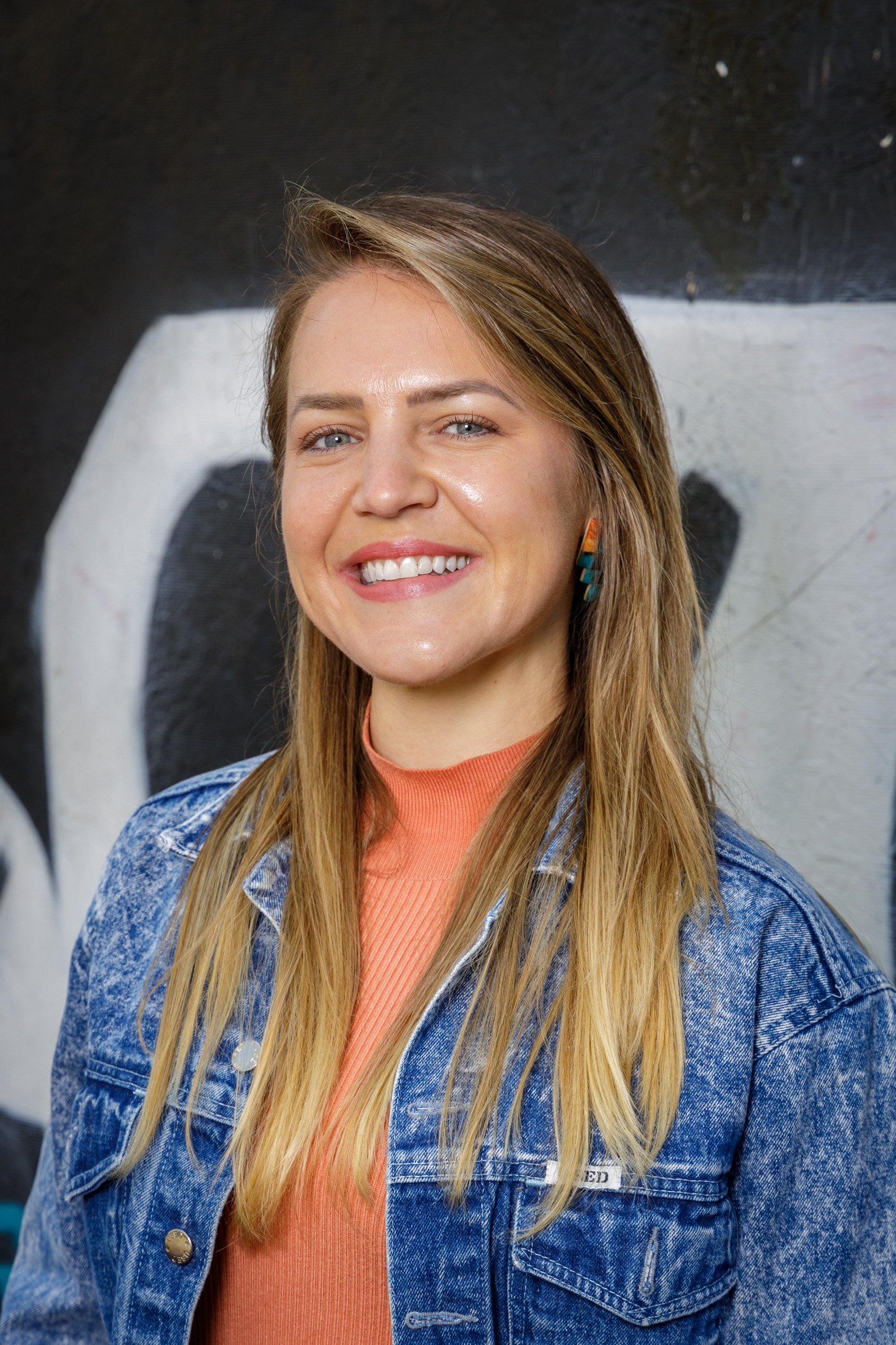 A woman with long blonde hair smiling, wearing a denim jacket and orange shirt, standing in front of a black and white painted background.