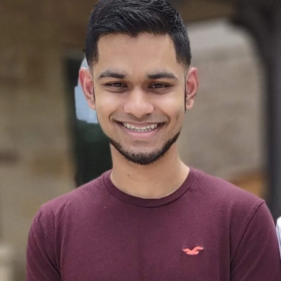 A young man with short dark hair and a beard, smiling, wearing a maroon t-shirt with a small logo, outdoors with blurred background.
