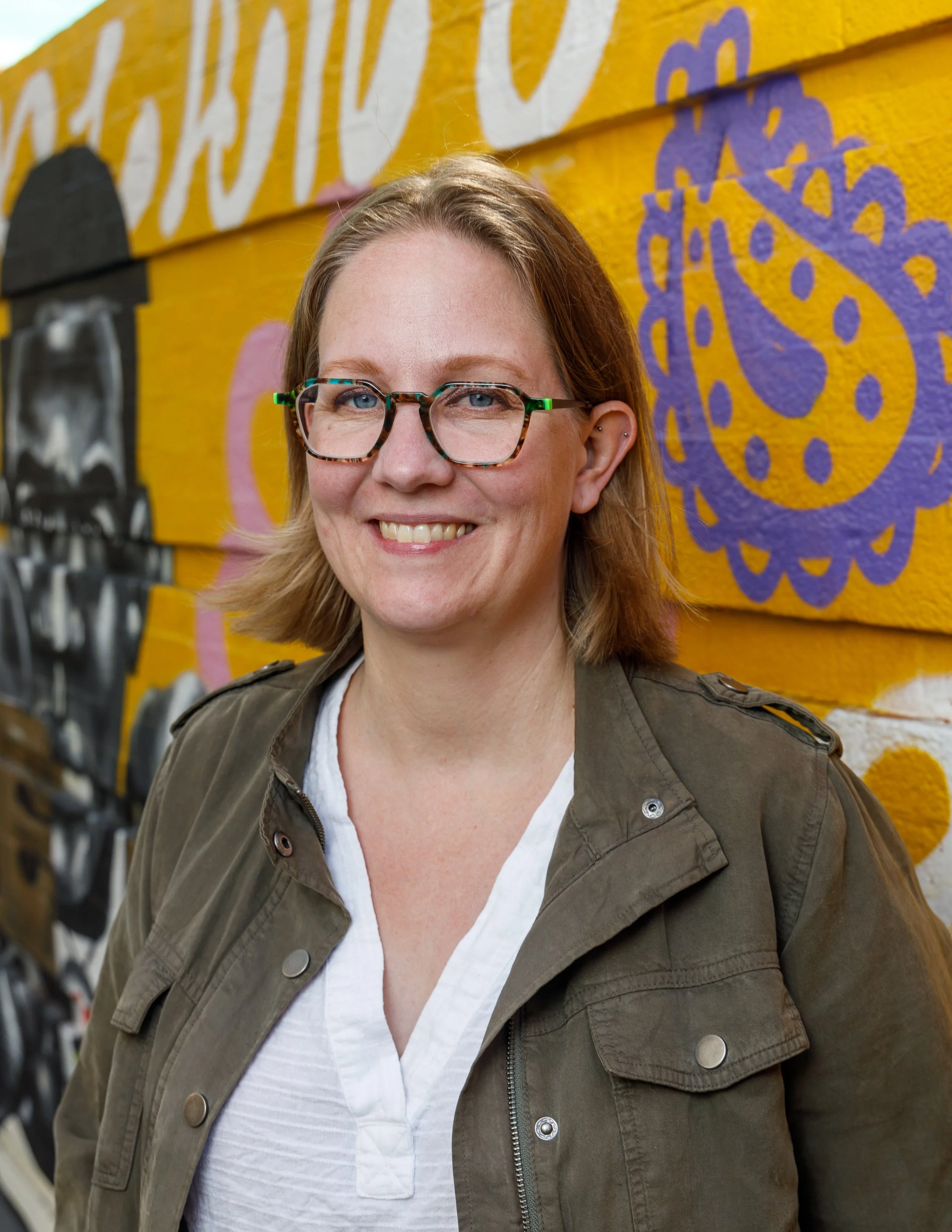 A woman with shoulder-length blonde hair, glasses, and a friendly smile standing in front of a colorful, graffiti-style mural on a wooden wall.