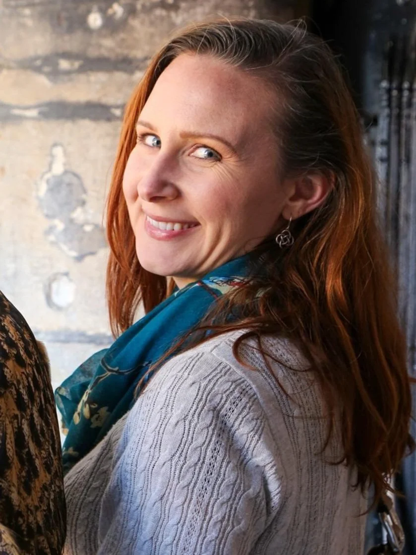 A woman with long red hair and blue eyes, smiling at the camera, wearing earrings, a patterned scarf, and a light-colored sweater, standing in front of a textured wall.