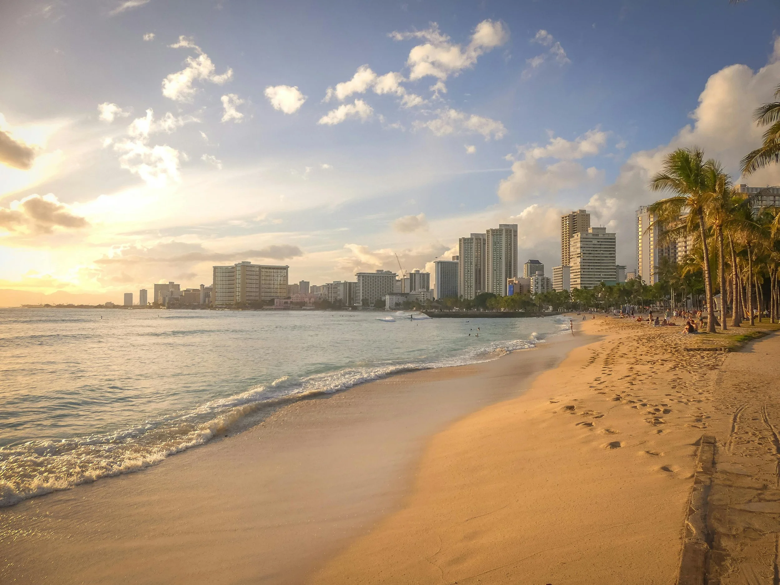 A sandy beach at sunset with a city skyline in the background, including tall buildings and palm trees along the shoreline.