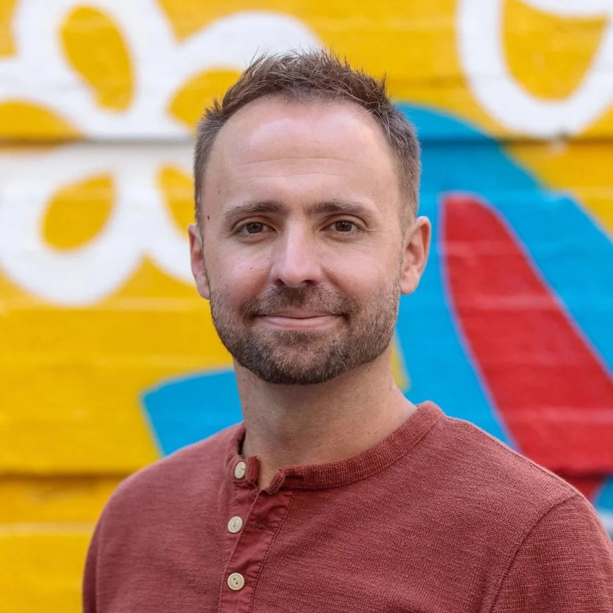 A smiling man with short brown hair and a beard, wearing a reddish-brown henley shirt, standing in front of a colorful mural with yellow, white, blue, and red designs.