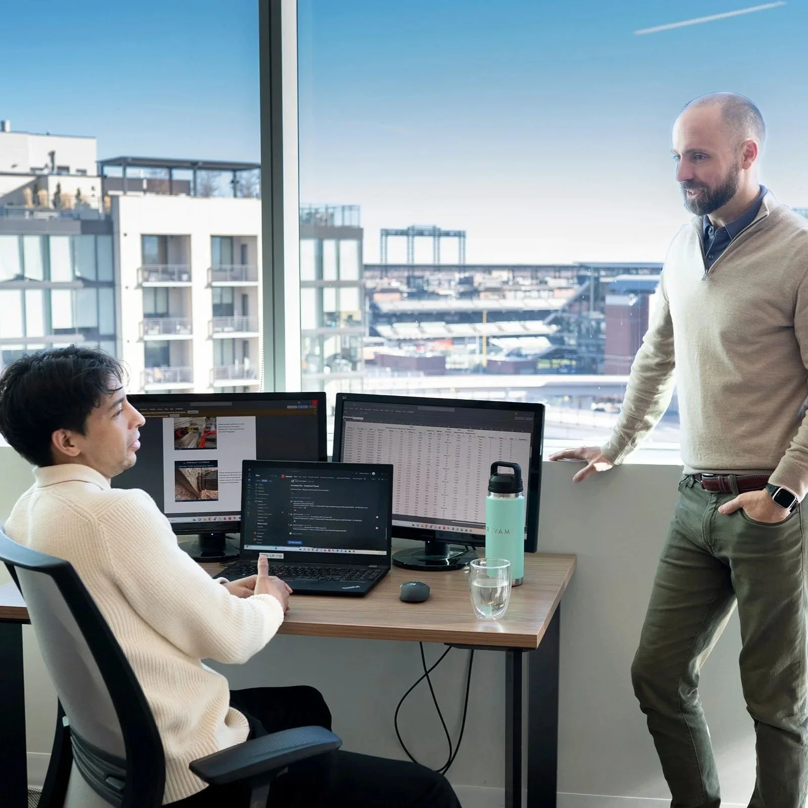 Two men in an office with multiple monitors, one sitting at the desk and the other standing, having a discussion. Large windows show a cityscape in the background.