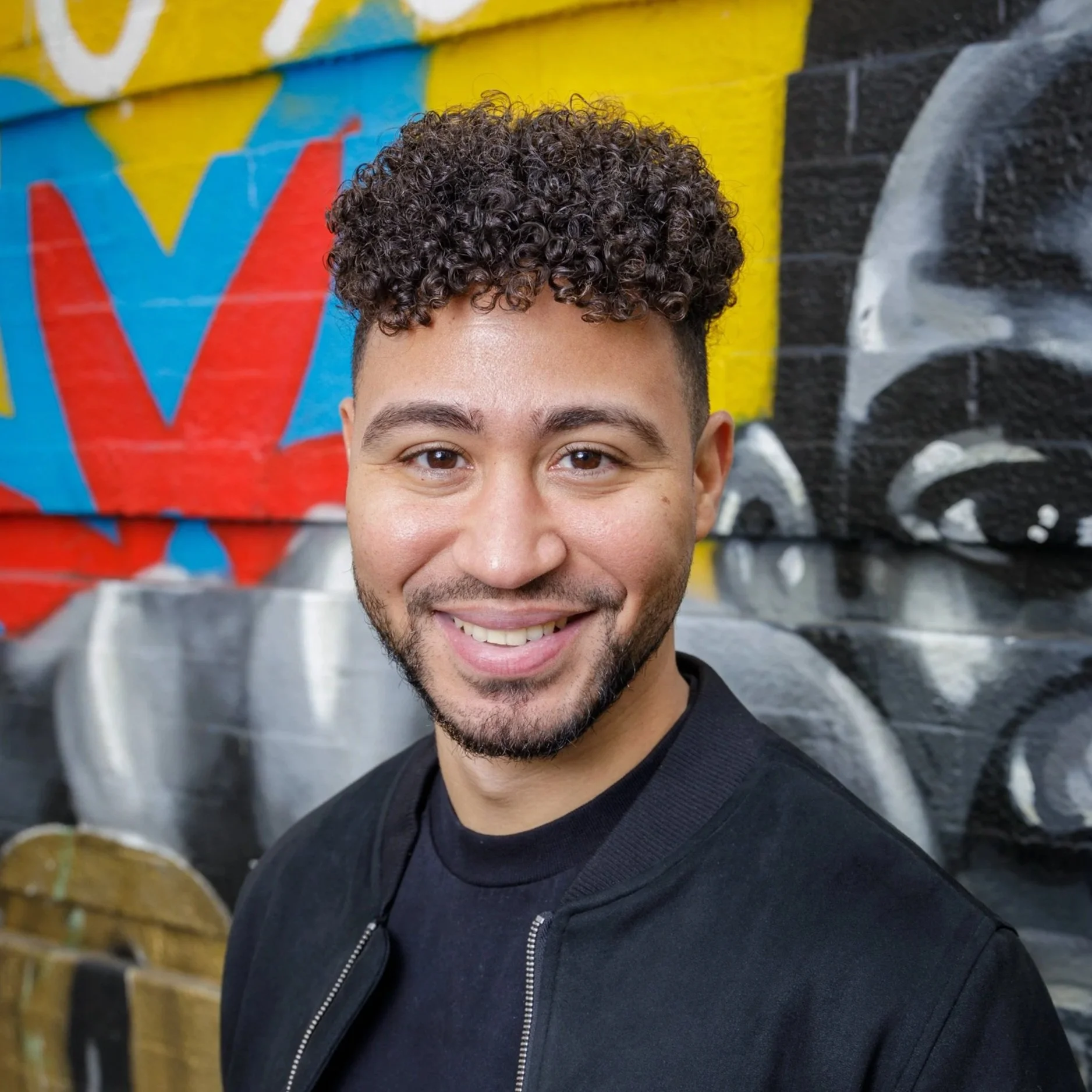 A young man with curly hair and a beard smiling, standing in front of a colorful graffiti wall.
