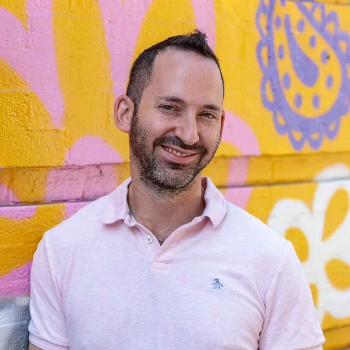A smiling man with short dark hair and a beard, wearing a light pink polo shirt, standing in front of a colorful mural wall with pink, yellow, and purple patterns.