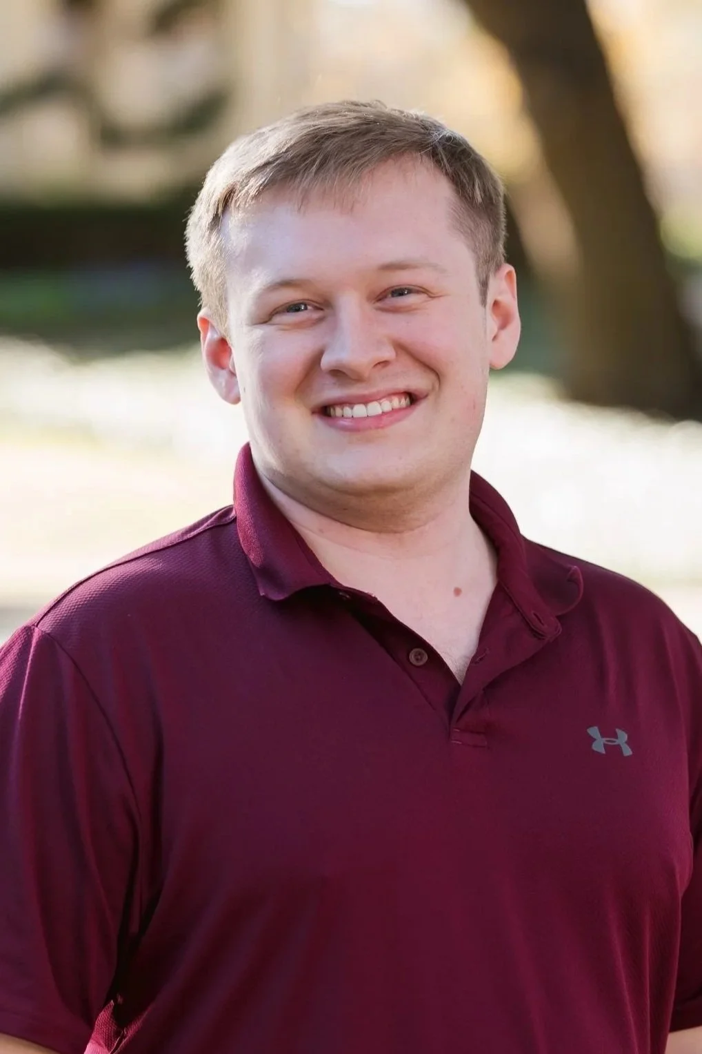 A smiling man with light skin, short blond hair, wearing a maroon polo shirt outdoors with blurred trees in the background.