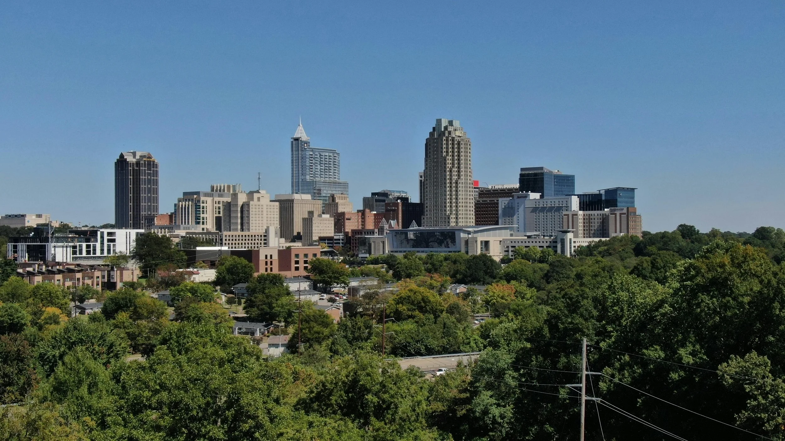 City skyline with tall buildings against a clear blue sky, with a foreground of green trees and residential houses.