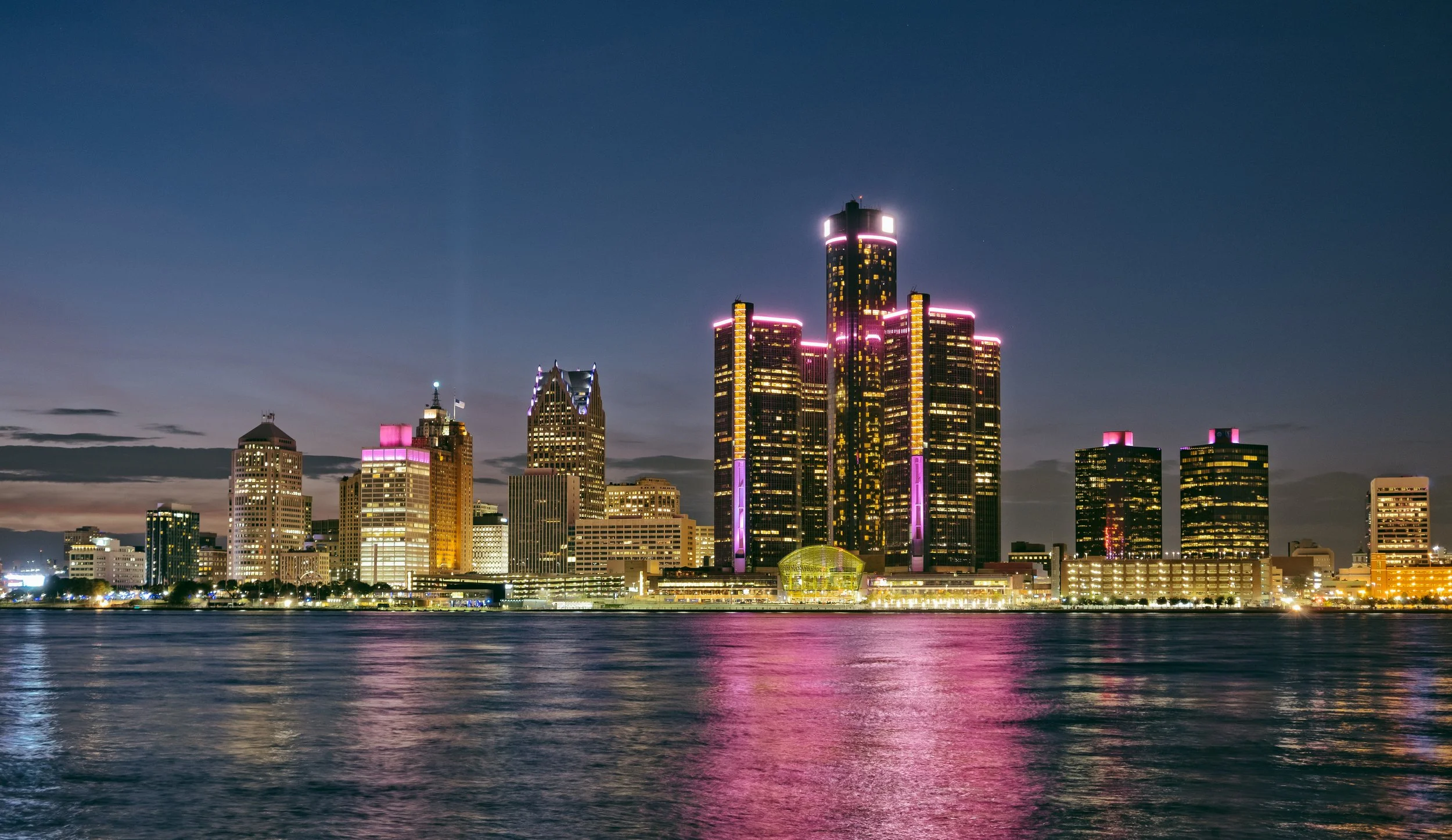 Night view of a city skyline with tall buildings illuminated by pink and yellow lights, reflecting on the water in the foreground.