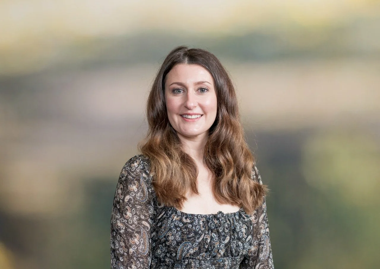 A woman with long wavy brown hair and blue eyes, smiling, outdoors with blurred trees in the background Revamp