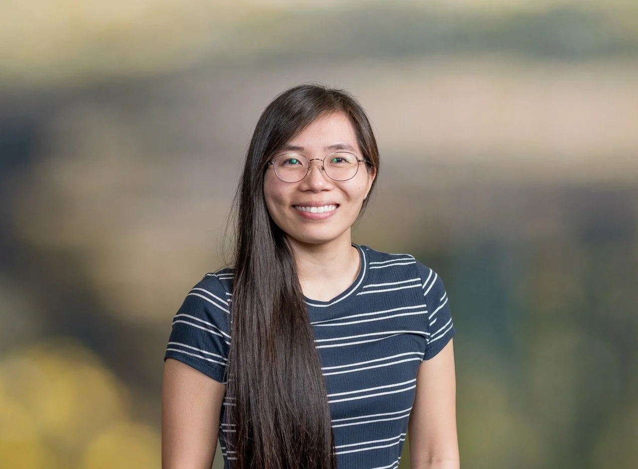 A young woman with long dark hair, wearing glasses and a black coat, smiling outdoors with autumn trees and a body of water in the background on a clear day.