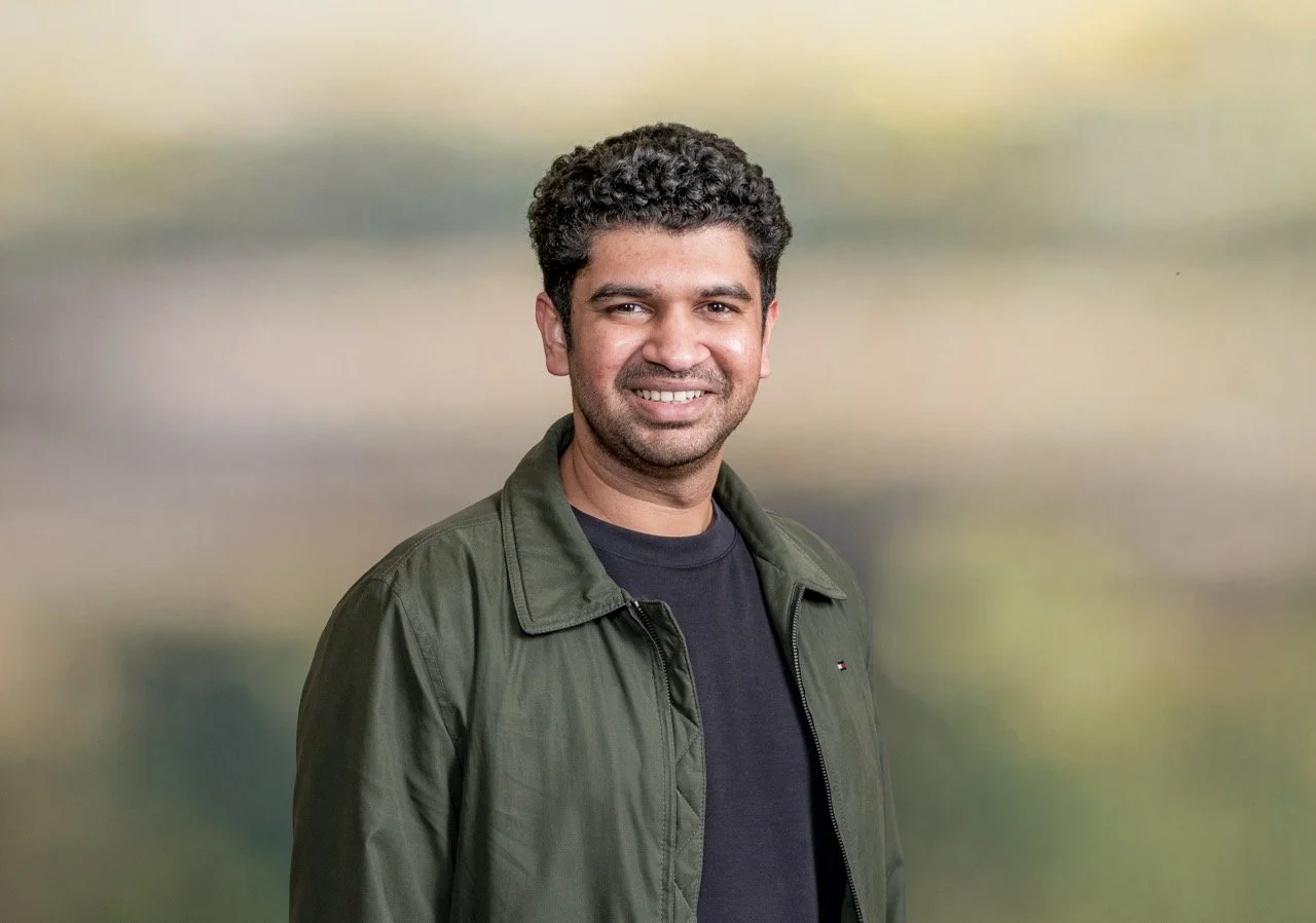A smiling young man with curly black hair, wearing a red shirt, standing against a plain light-colored background.