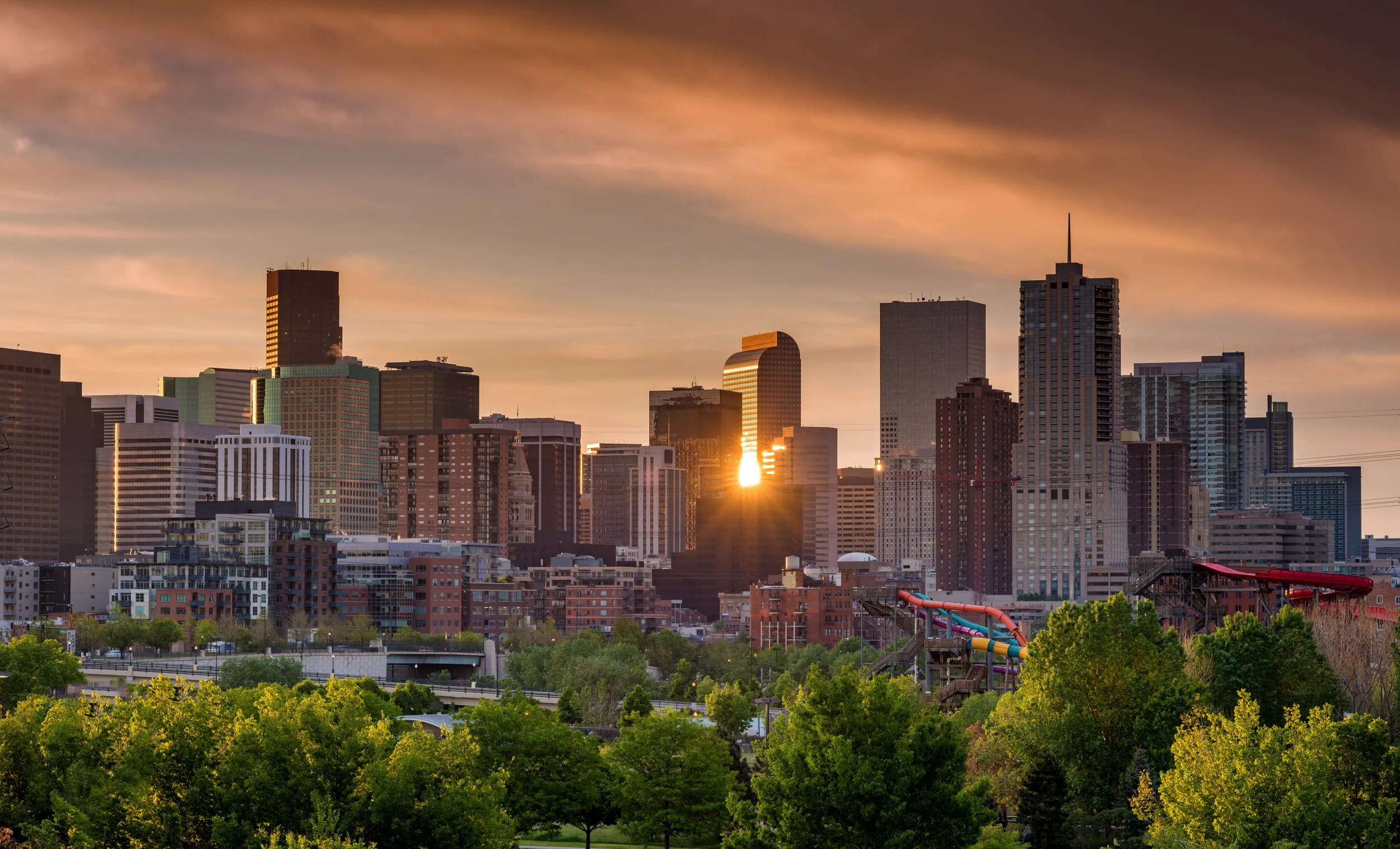 Sunset over a city skyline with green trees in the foreground and colorful water slides.