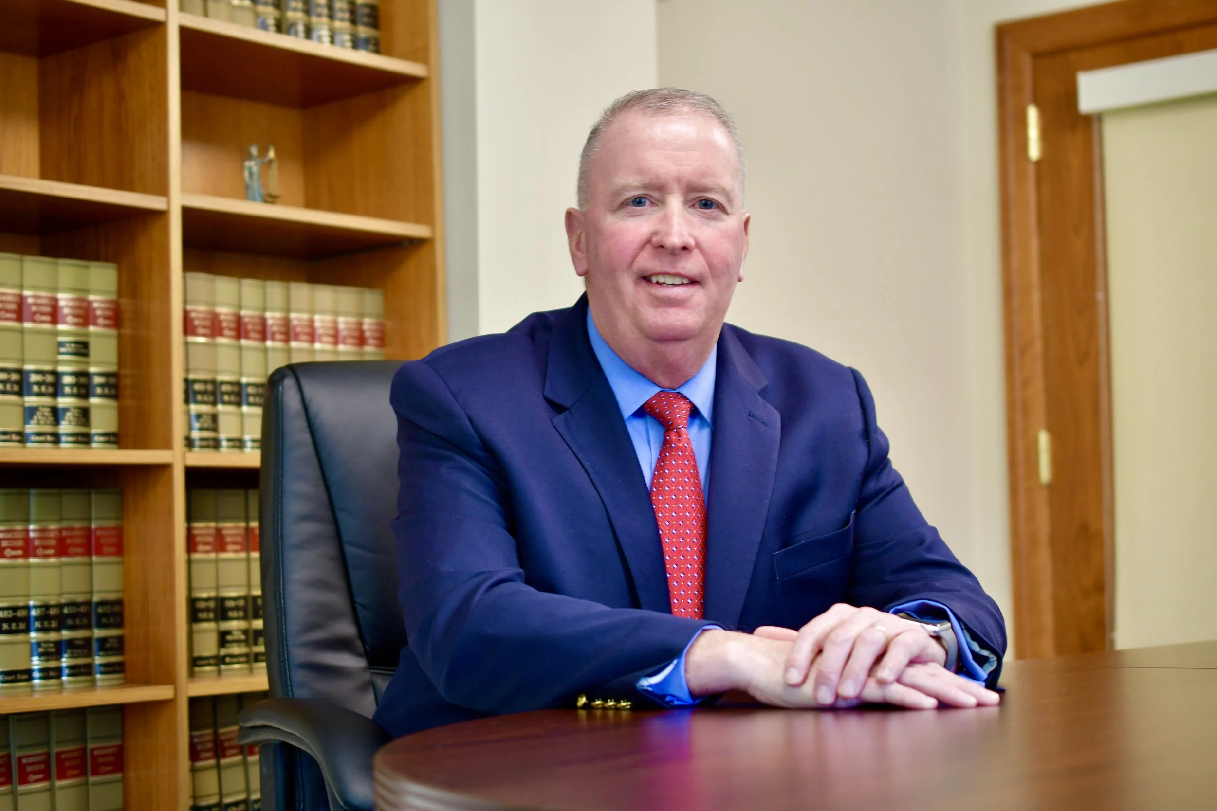 A man in a dark blue suit and red polka dot tie sitting at a wooden desk in an office with wooden bookshelves and legal books in the background.