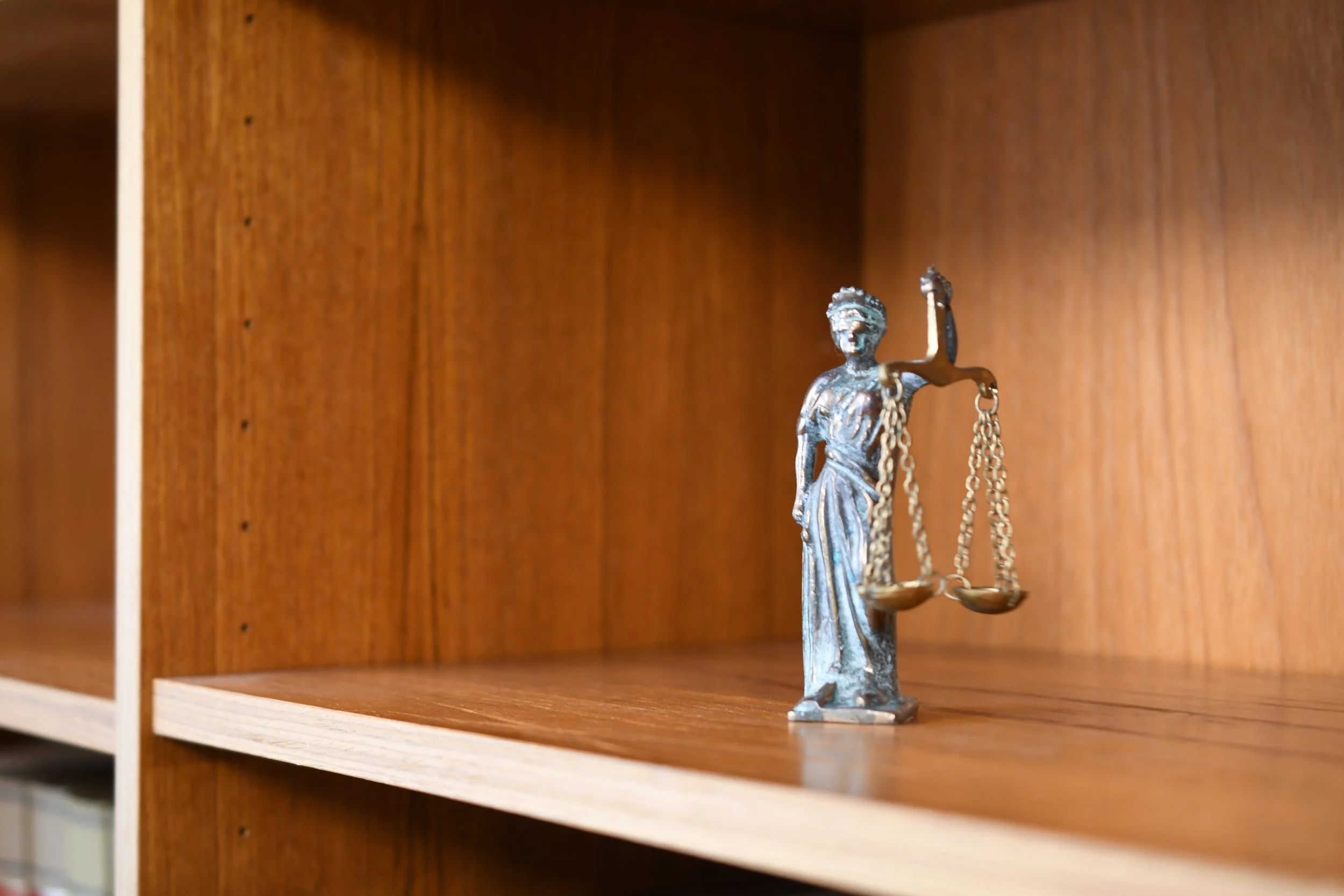 Small metallic Lady Justice statue holding scales, placed on a wooden shelf against a wooden wall.