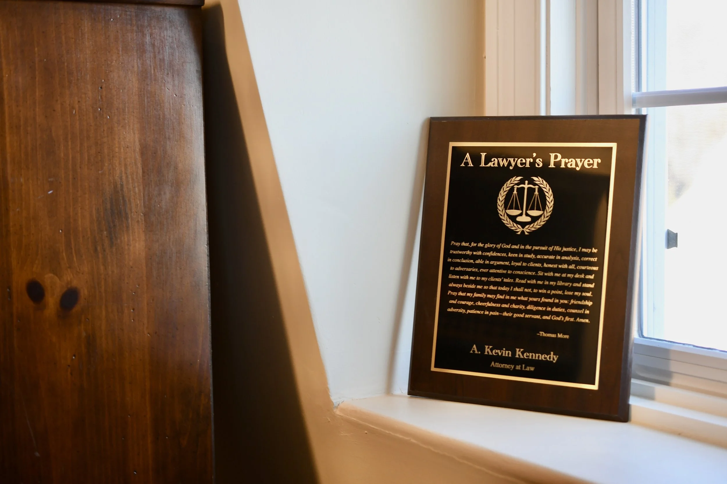 A framed black plaque titled "A Lawyer's Prayer" sitting on a windowsill next to a window with blinds. The plaque features an emblem of scales inside a laurel wreath and a prayer text below.