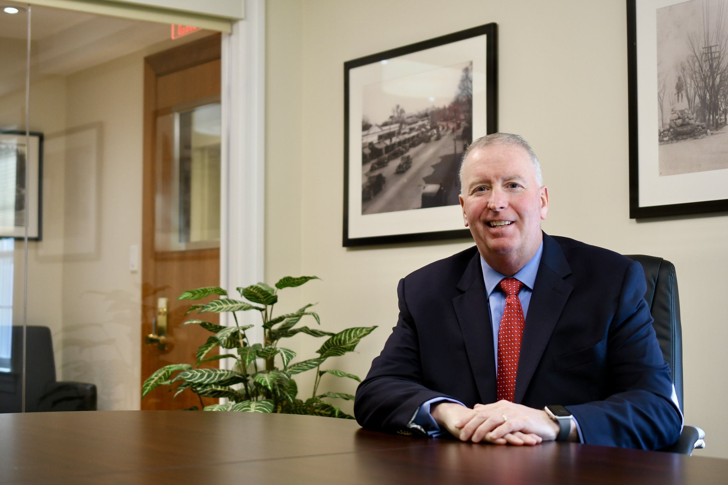 A smiling middle-aged man in a business suit and red tie sitting at a wooden conference table in an office, with framed photographs and a plant on the wall behind him.