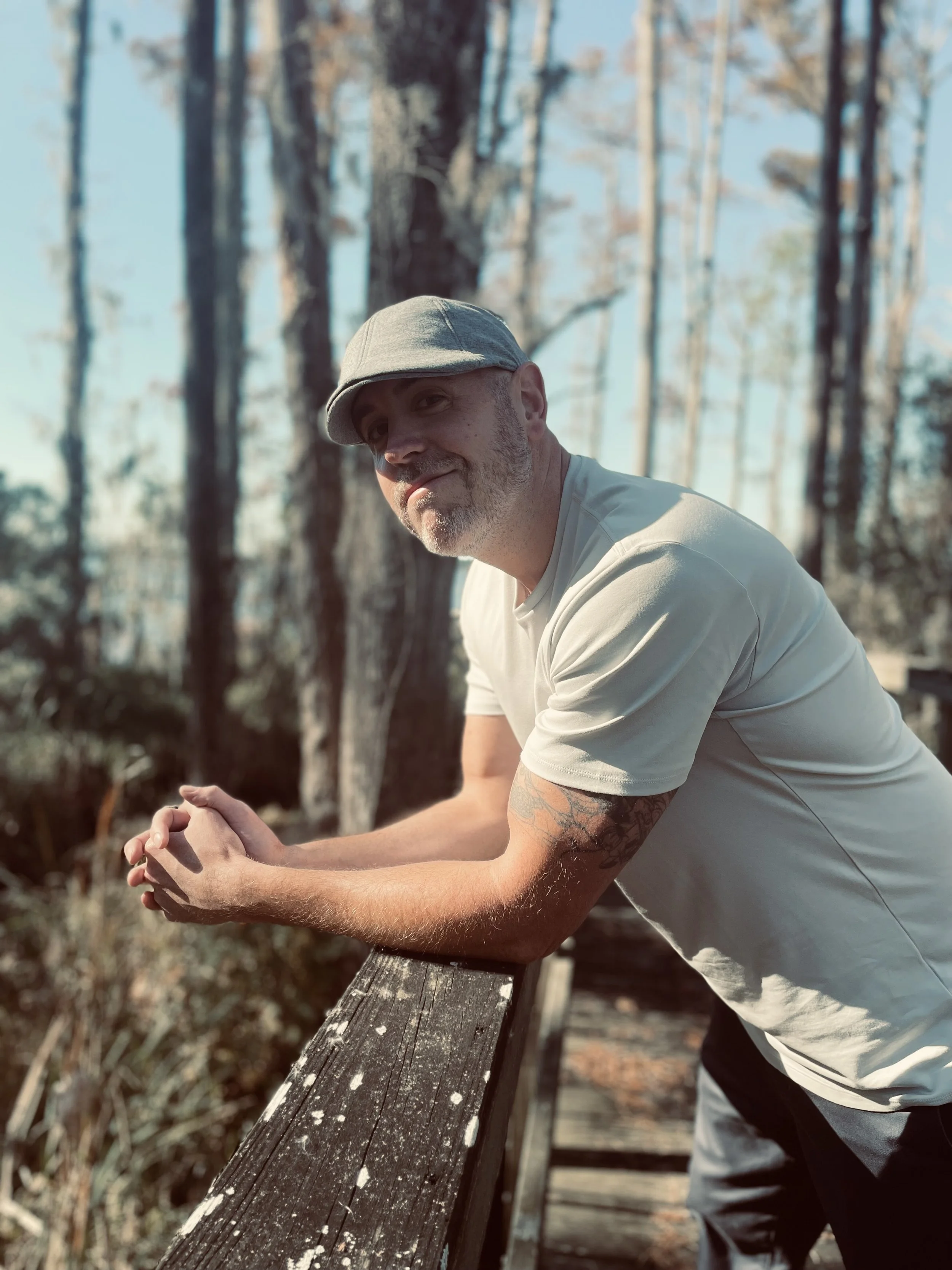 A man with a beard, wearing a gray cap and a white t-shirt, leans on a wooden railing outdoors in a wooded area, with tall trees and a blue sky in the background.