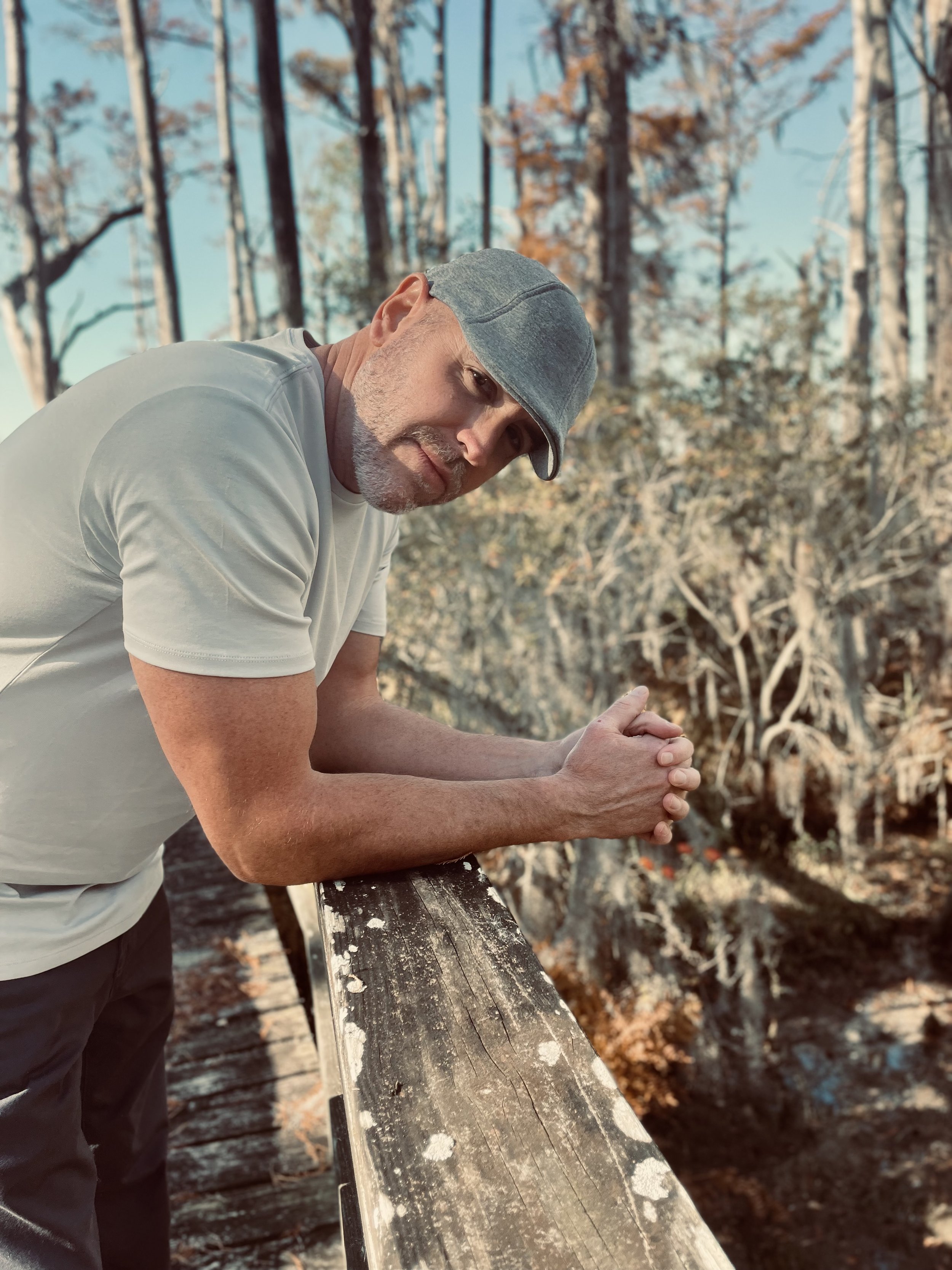 A man with a beard, wearing a gray cap and light-colored t-shirt, leaning on a weathered wooden railing outdoors in a wooded area during daytime.