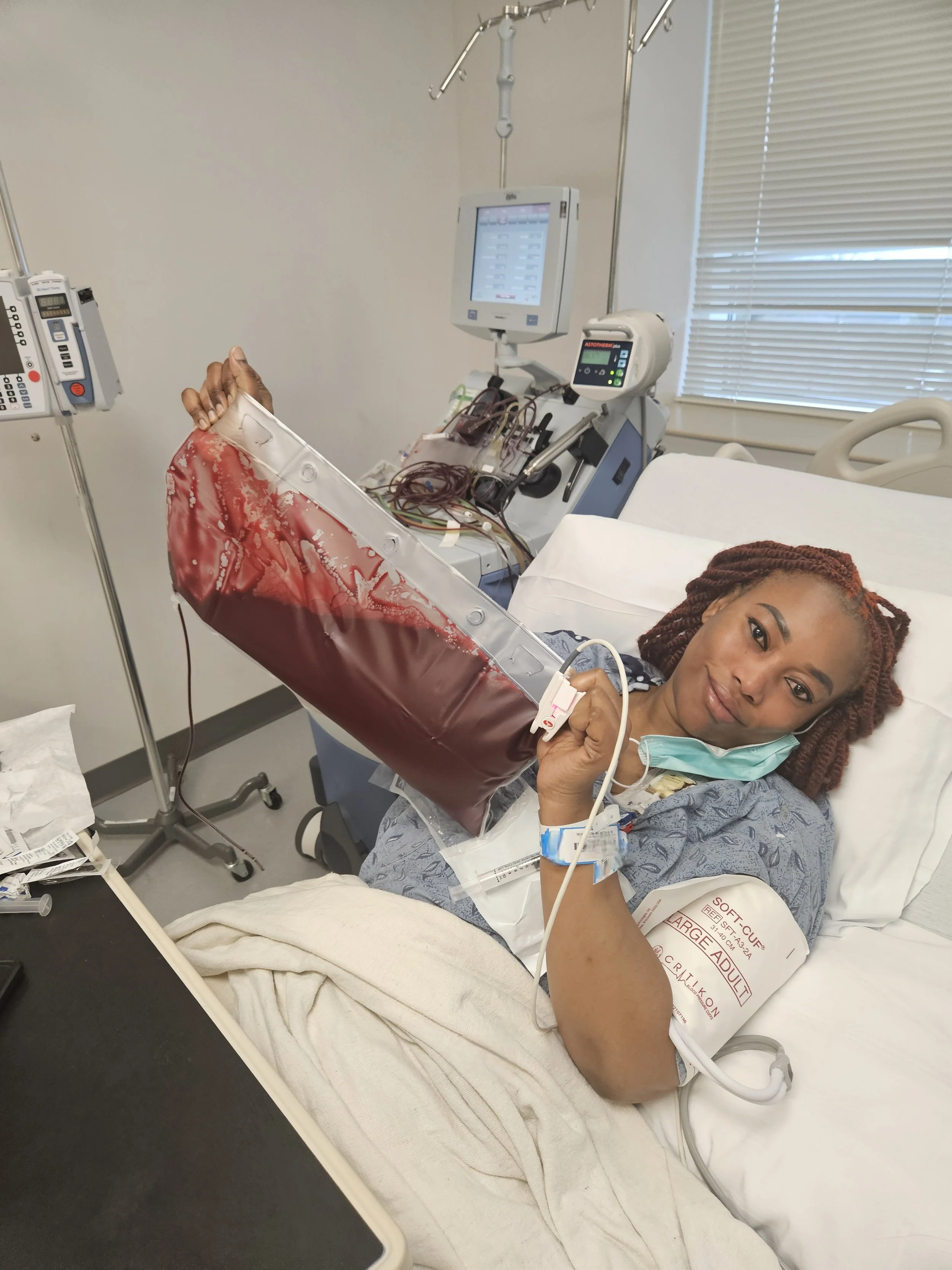 A woman lying in a hospital bed holding a blood bag, smiling at the camera, with medical equipment and monitors around her.