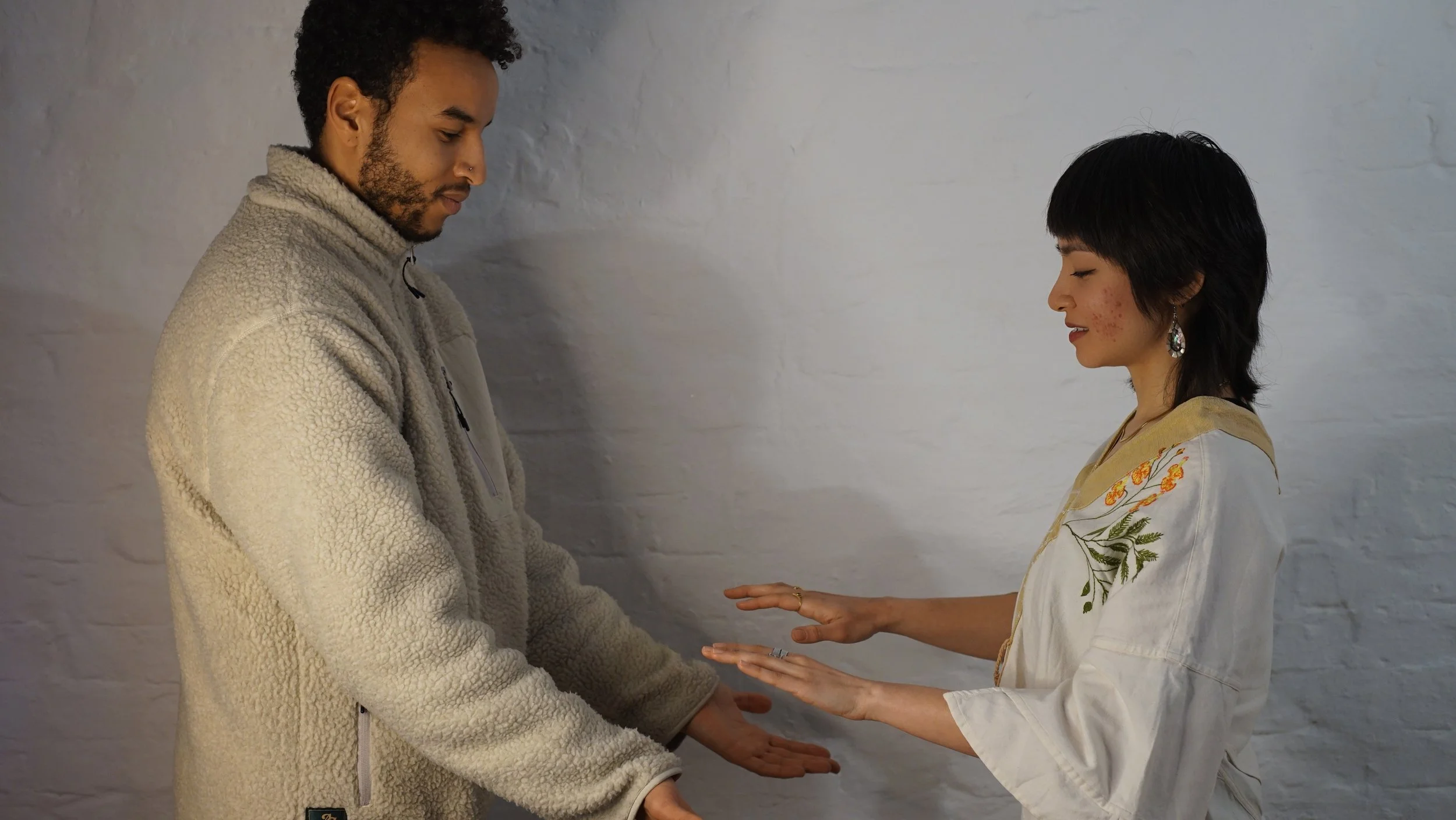 Two people standing facing each other, holding hands, with eyes closed and appearing to be engaged in a mindful or spiritual practice against a plain white brick wall.