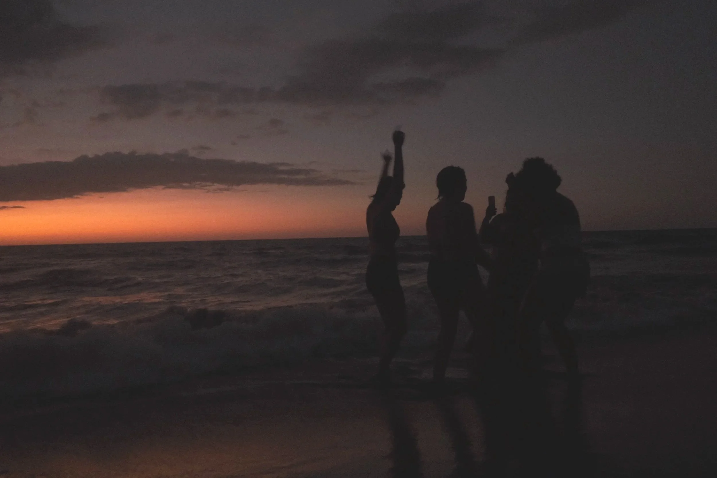 Silhouettes of three people on a beach at sunset, with clouds and a colorful sky in the background.