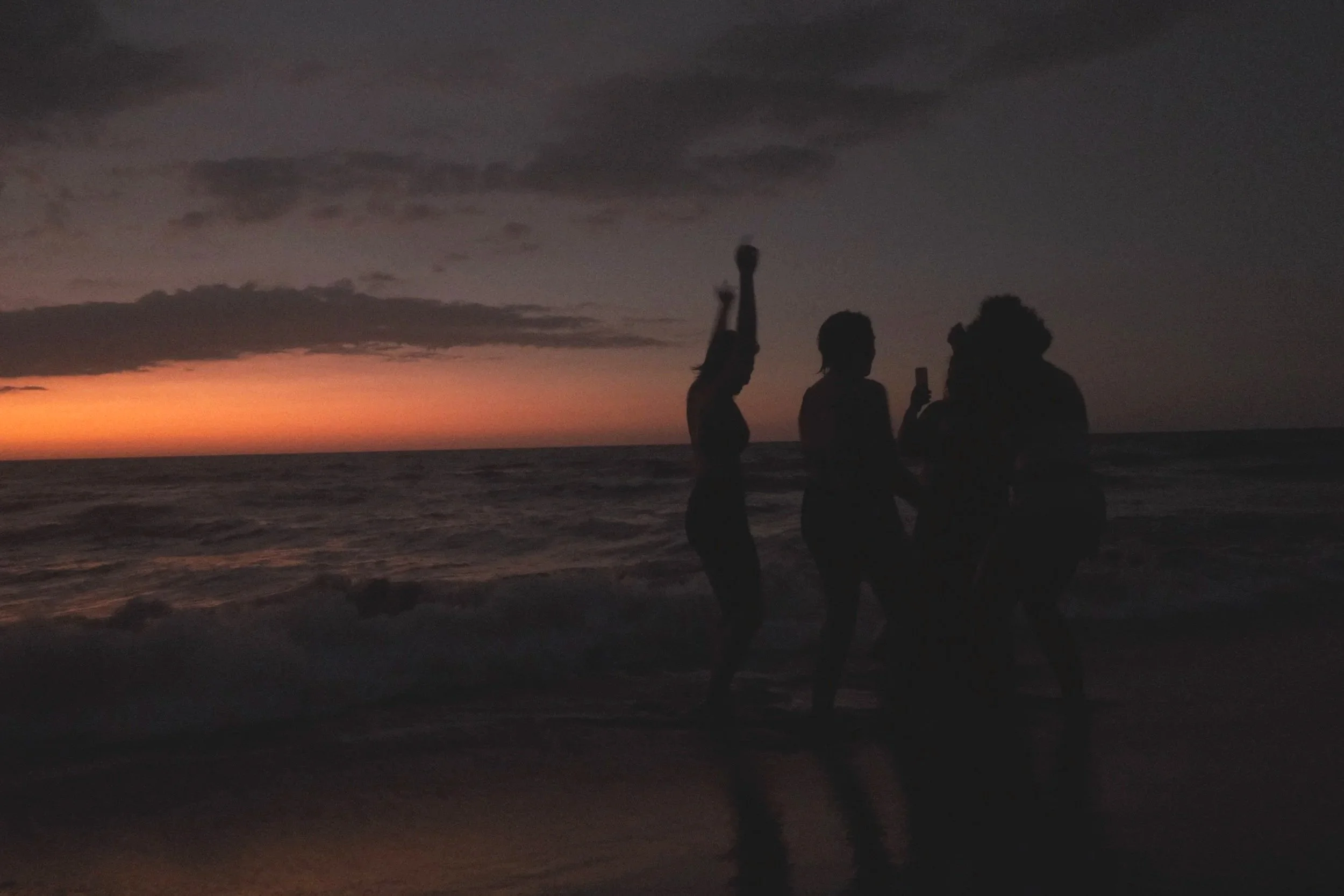 Silhouettes of four people standing on a beach at sunset, with dark clouds in the sky and ocean waves in the background.