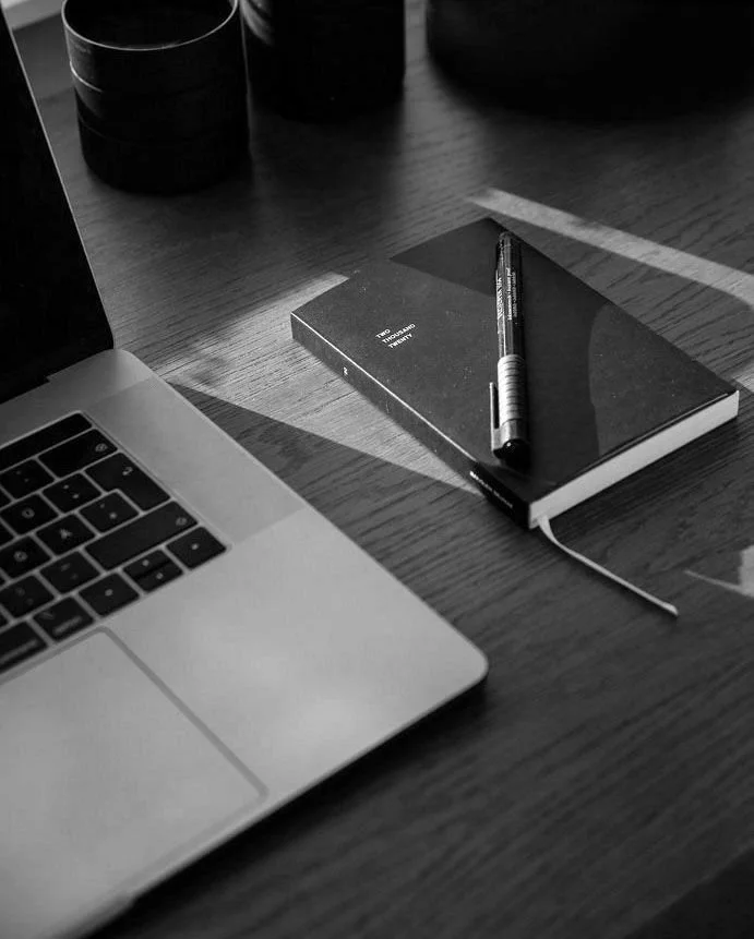 Close-up of a laptop, a closed notebook with a pen on top, a cup, and part of a table in black and white.