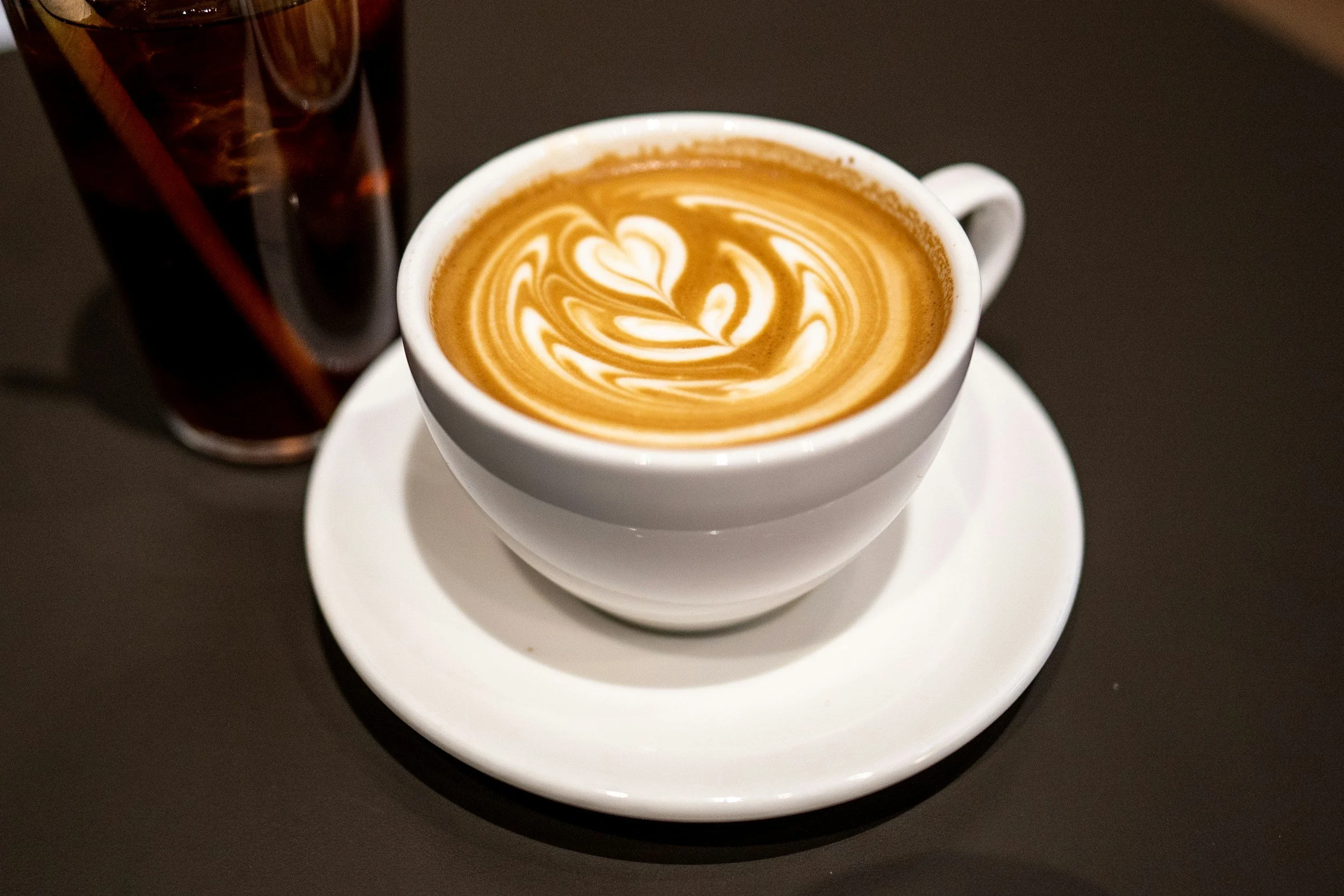 Cup of latte with latte art on top, placed on a saucer, with a glass of iced coffee in the background.