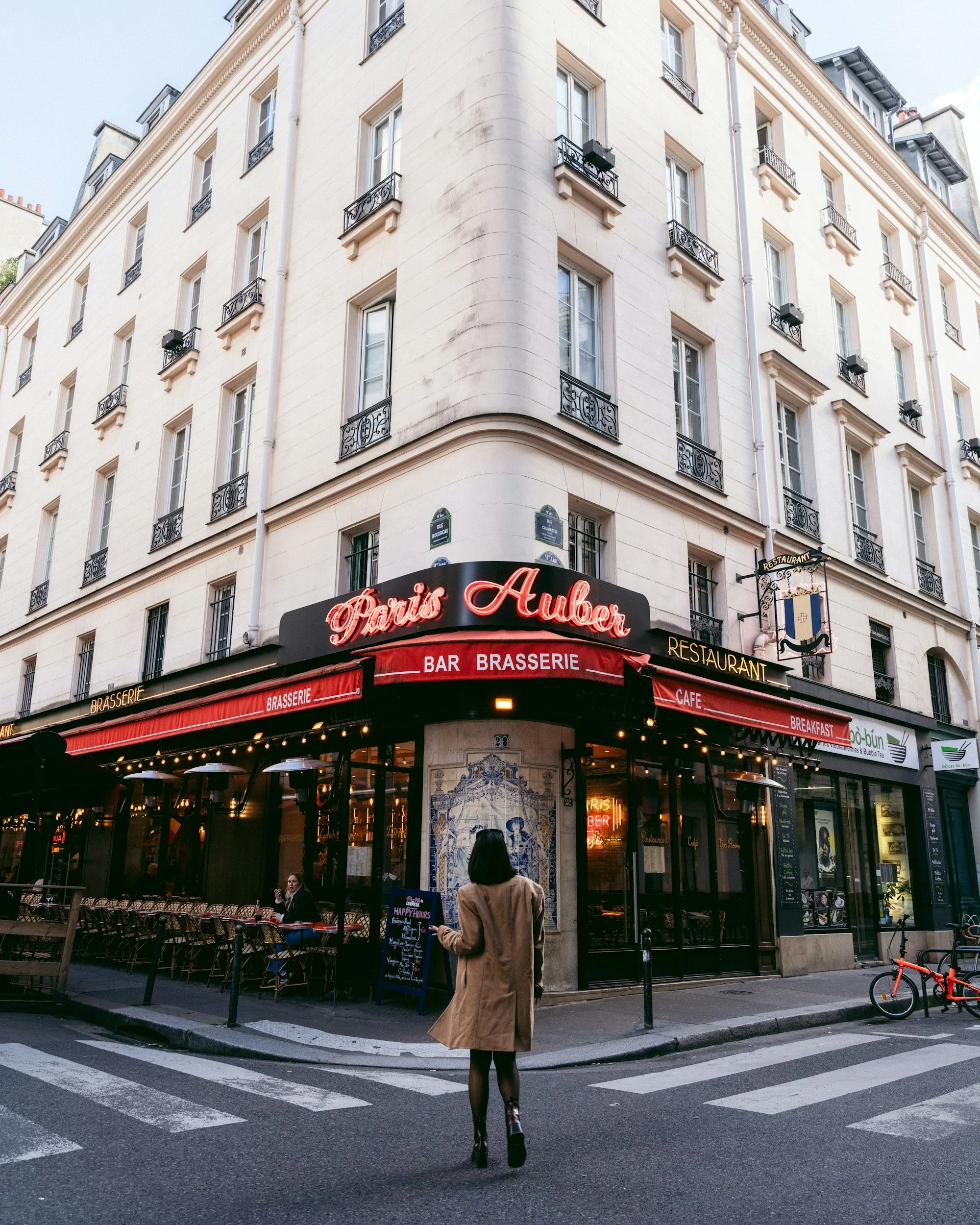 A woman crossing the street in front of a Parisian restaurant named 'Paris Auber' on a corner in Paris, France. The restaurant has outdoor seating and its signage includes 'Bar Brasserie,' 'Restaurant,' 'Cafe,' and 'Breakfast.'