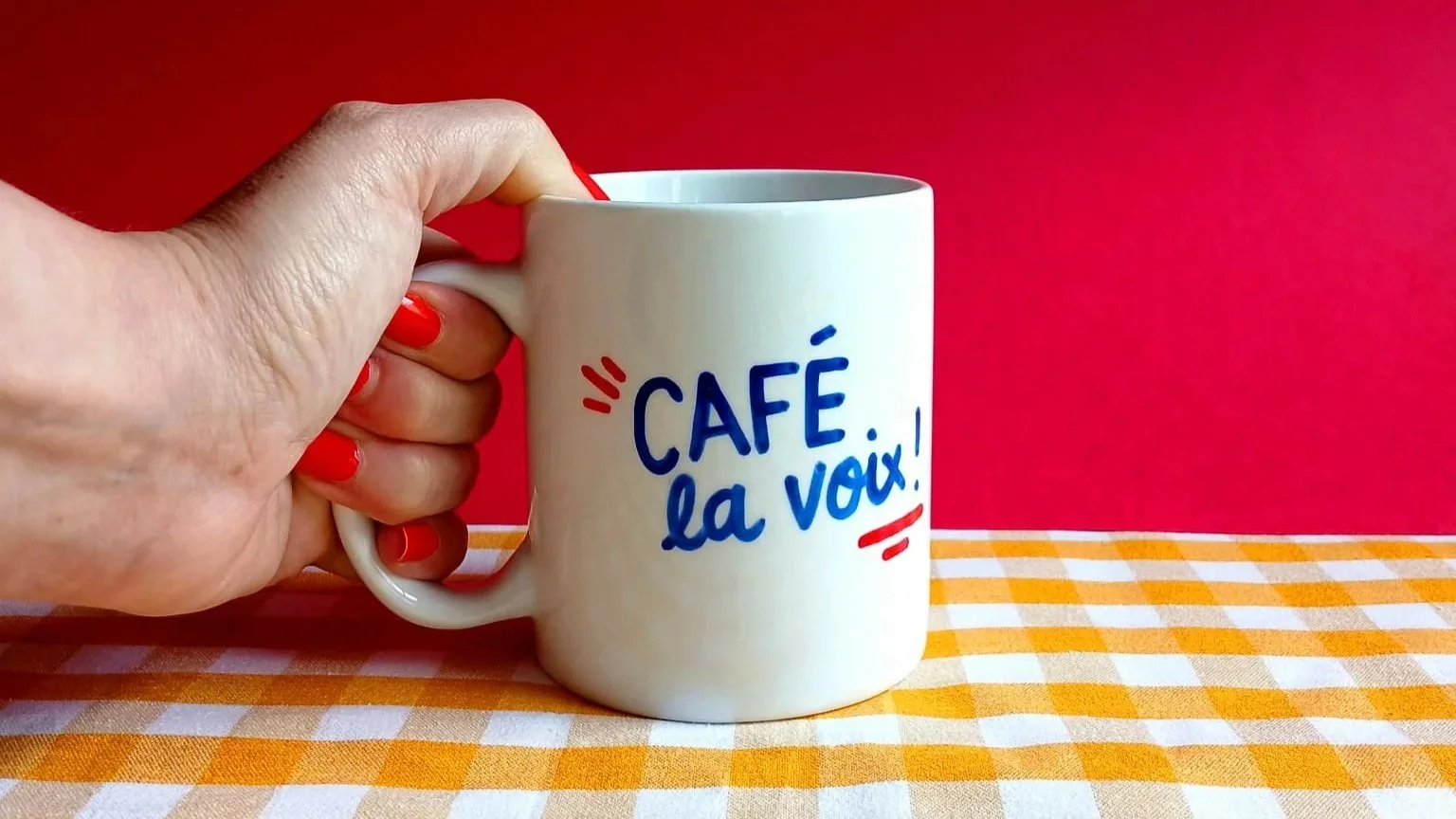 Tasse blanche avec le texte 'Café, la voix!' en bleu et rouge, tenue par une main avec ongles rouges, sur une nappe à carreaux orange et blanc, fond rouge.