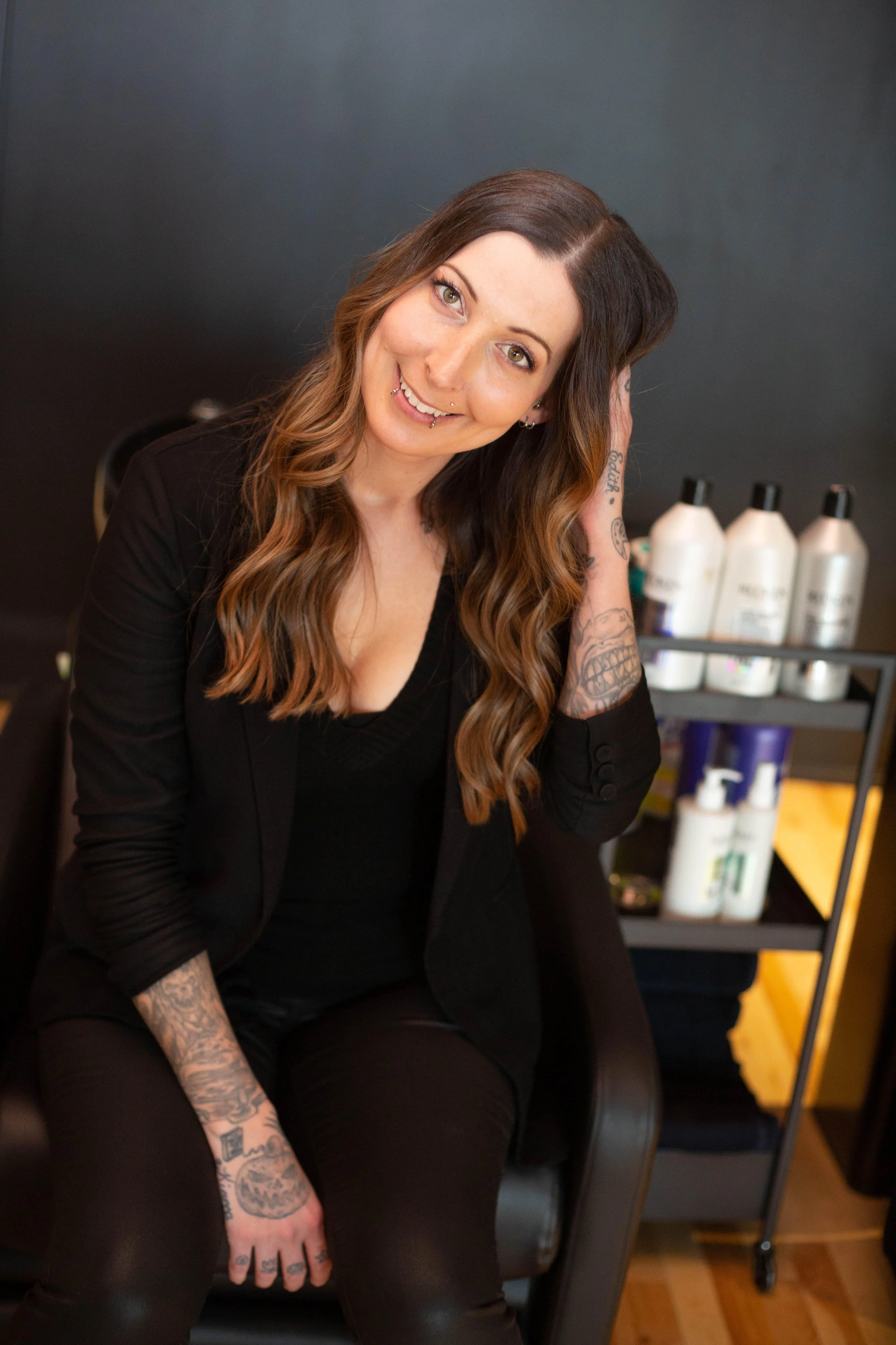 A woman with long, wavy brown hair, tattoos on her arms, and piercings on her lips. She is sitting in a black salon chair, wearing a black blazer and black clothing, smiling at the camera. Behind her on a small trolley are bottles of hair products.