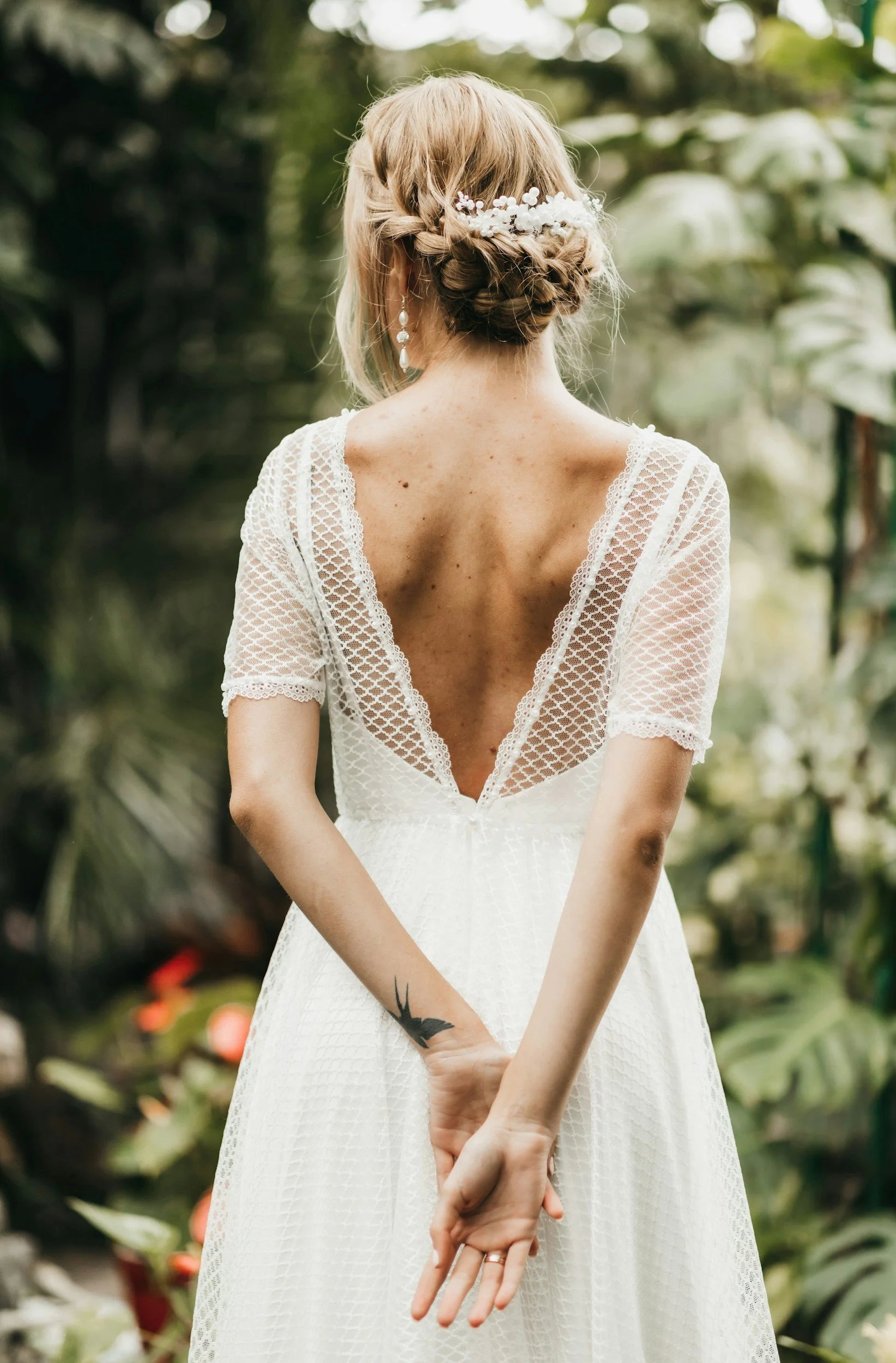 Woman in wedding dress with open back, braided hairstyle with floral hair accessory, standing outdoors in a lush, green garden.