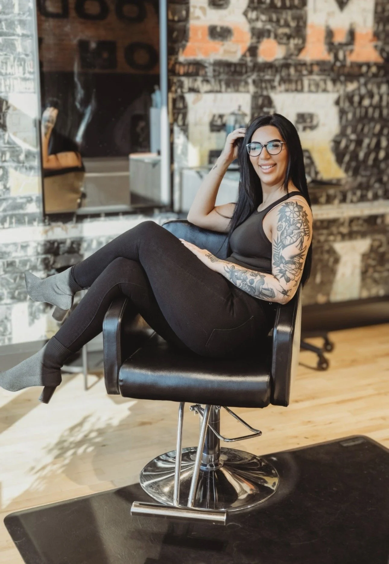 A woman with black hair, glasses, and tattoos on her arms, smiling and sitting on a salon chair in a tattoo parlor, with brick walls and mirrors in the background.