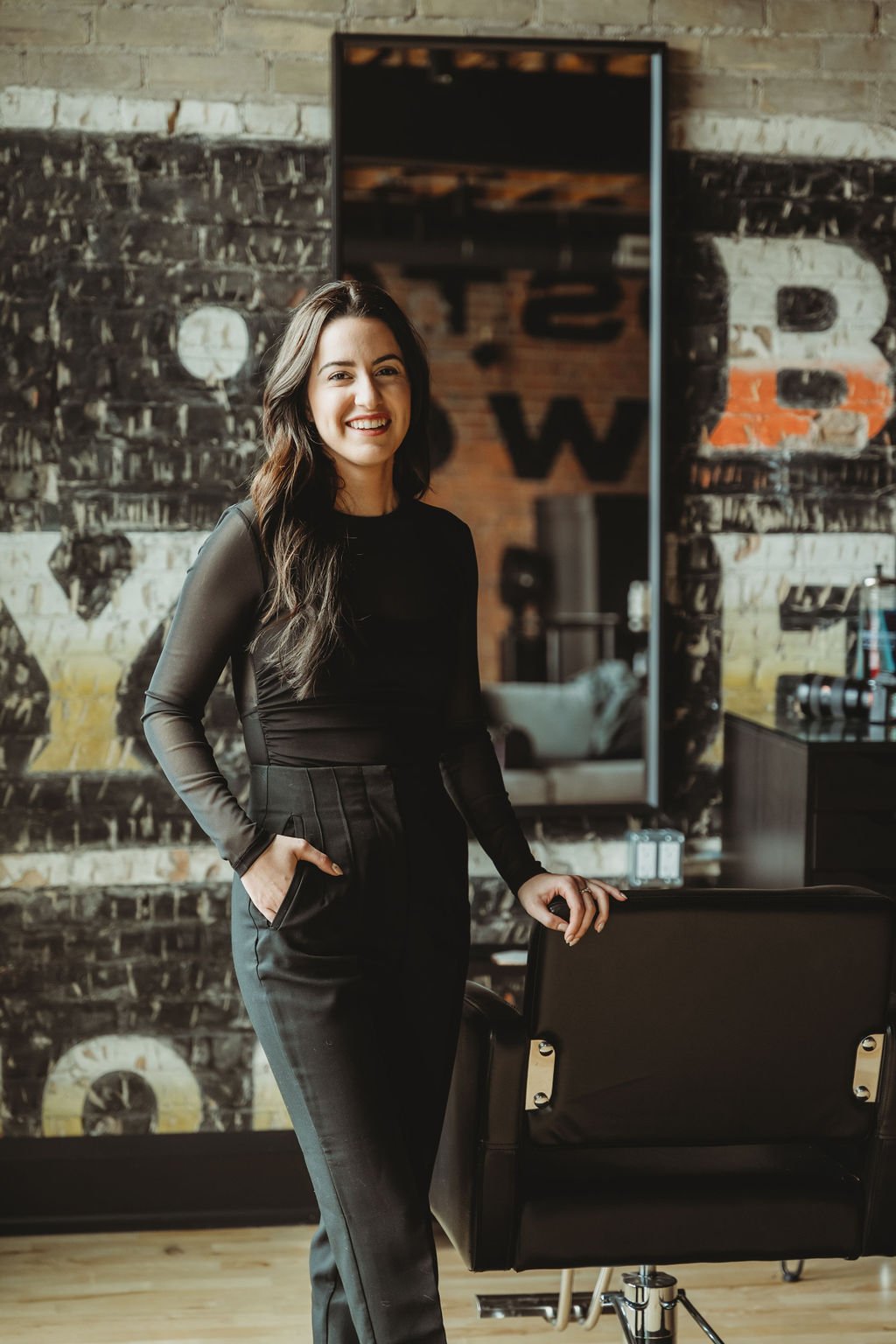 A woman with long dark hair, dressed in black, smiling and standing in a modern hair salon with exposed brick walls and a large mirror.