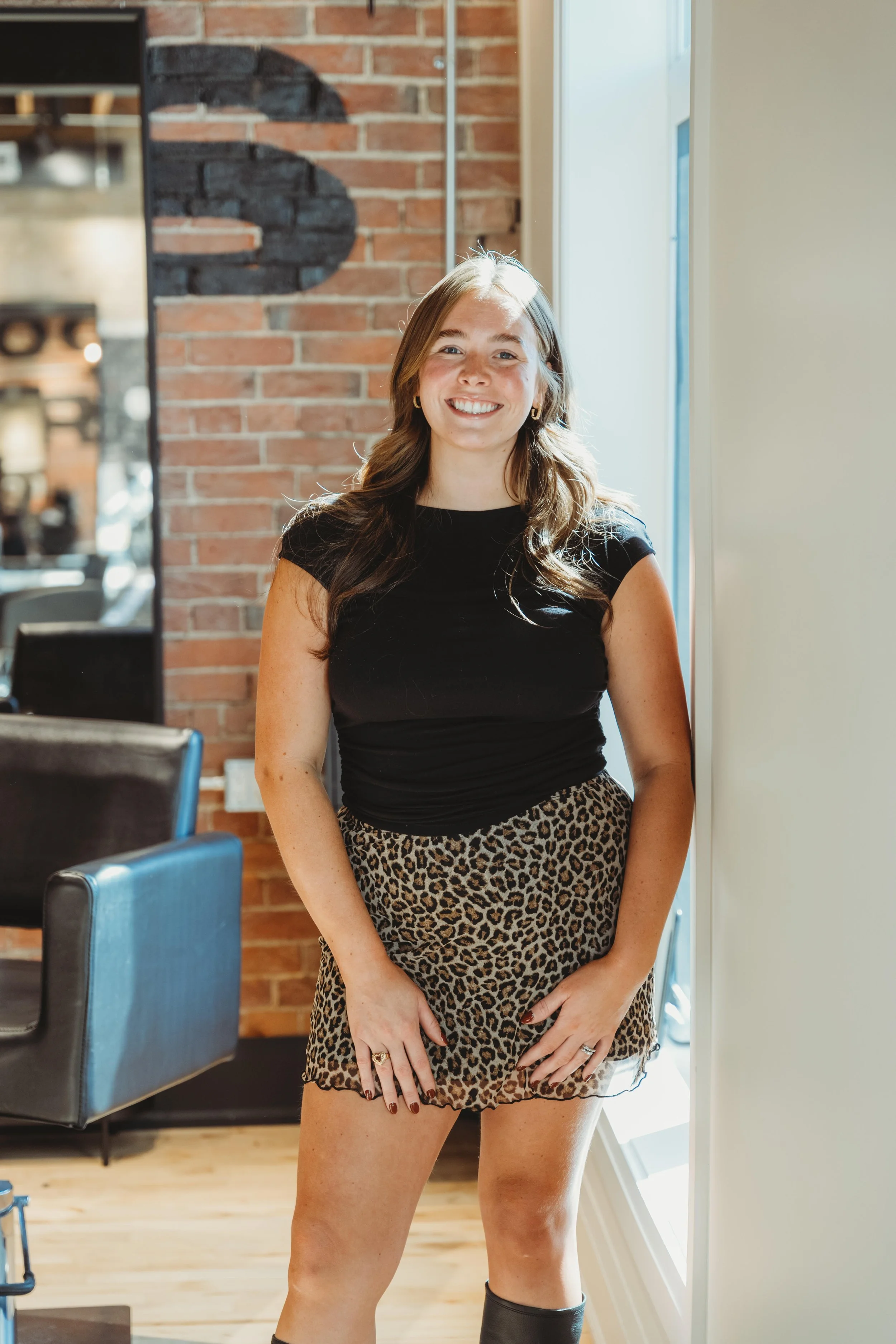 A young woman with long brown hair, wearing a black top and leopard print skirt, standing by a large window in a modern, industrial-style room and smiling at the camera.