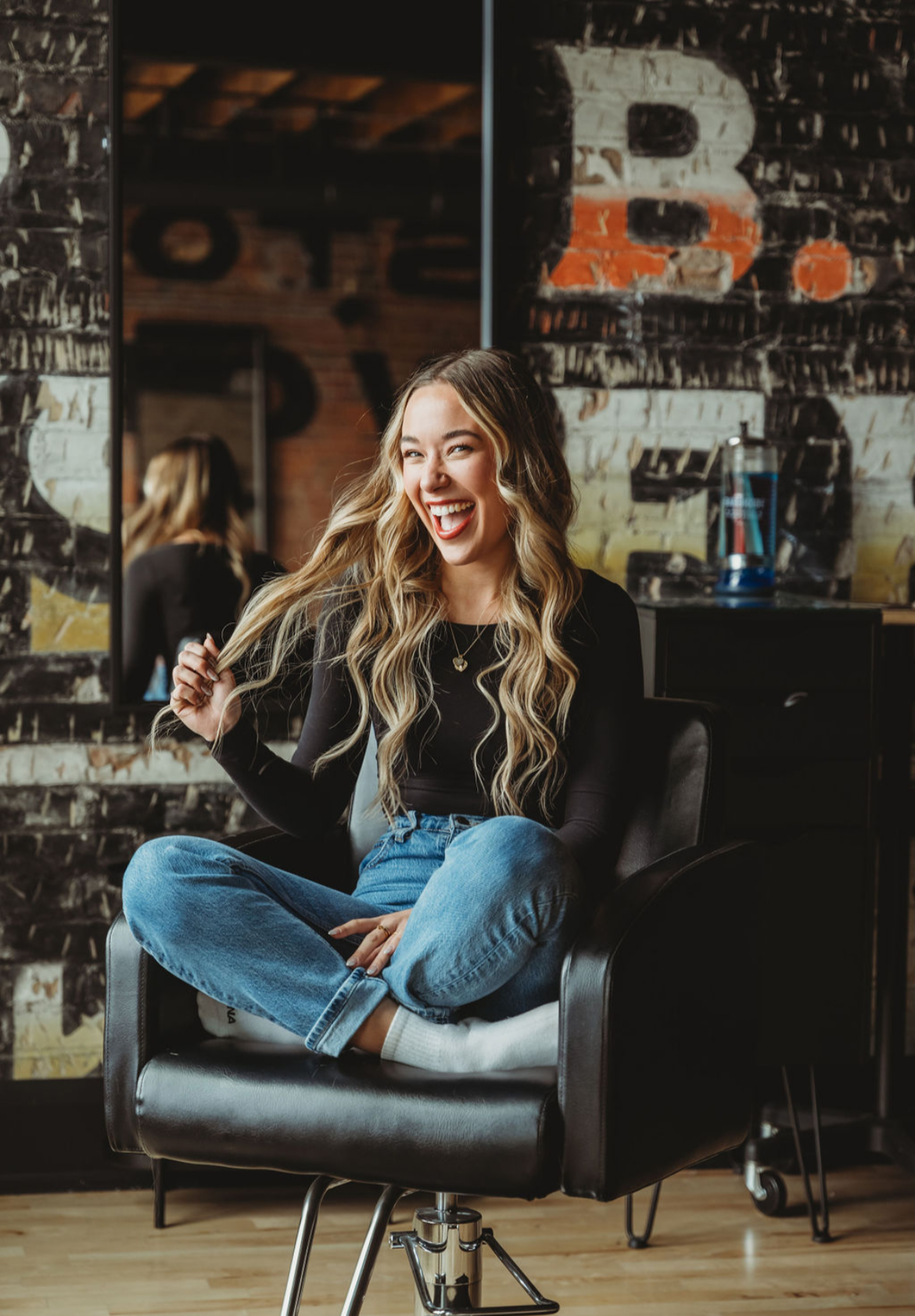 A woman with long, wavy blonde hair sits cross-legged and laughs while holding a strand of her hair in a salon chair. She wears a black long-sleeve top and blue jeans. The background features a graffiti-style brick wall and a mirror.