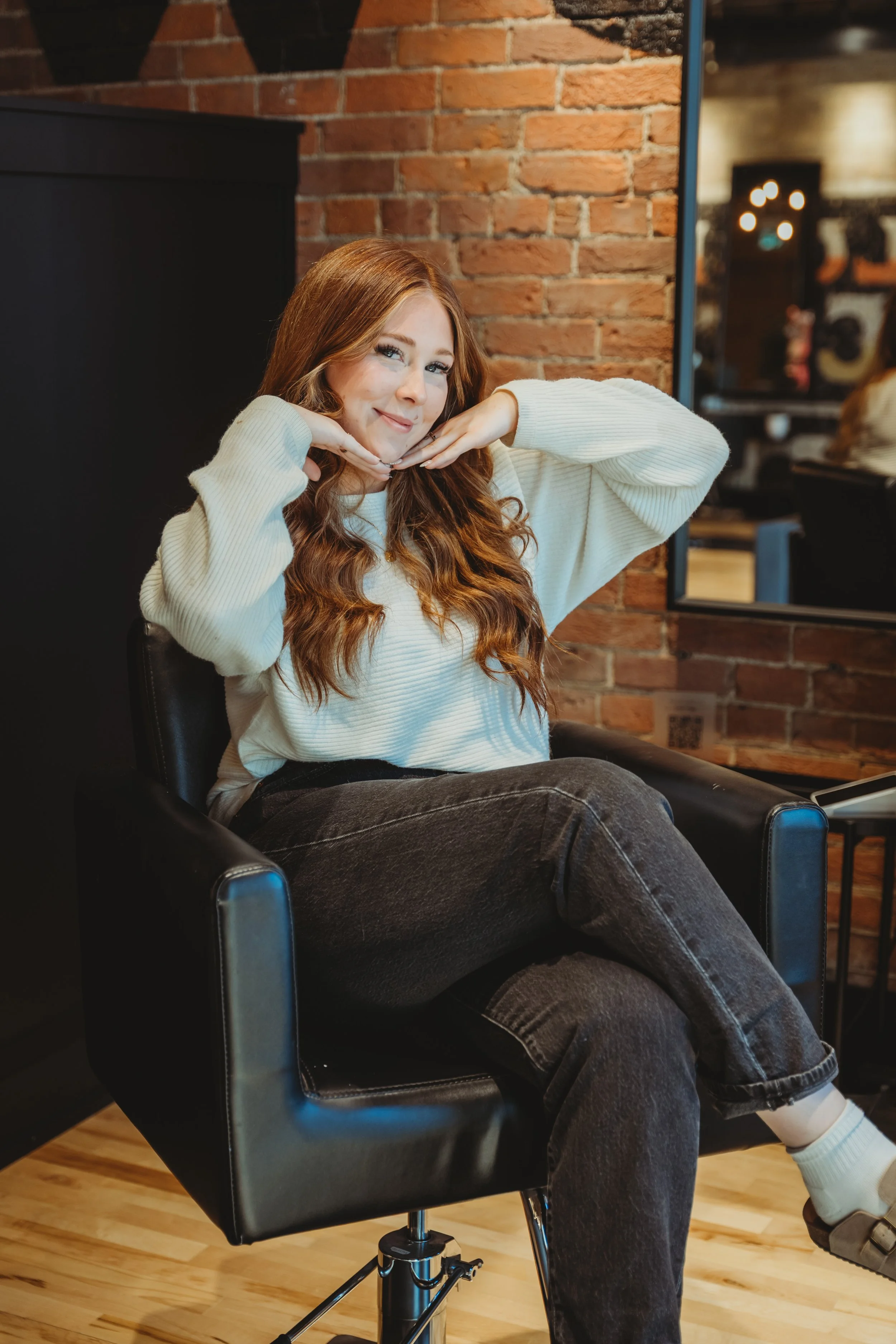 A young woman with long, wavy red hair sitting in a black salon chair in a salon with brick walls. She is smiling slightly and poses with her chin resting on her hands, elbows raised. She is wearing a white sweater, dark jeans, and gray slippers. A mirror is visible behind her.