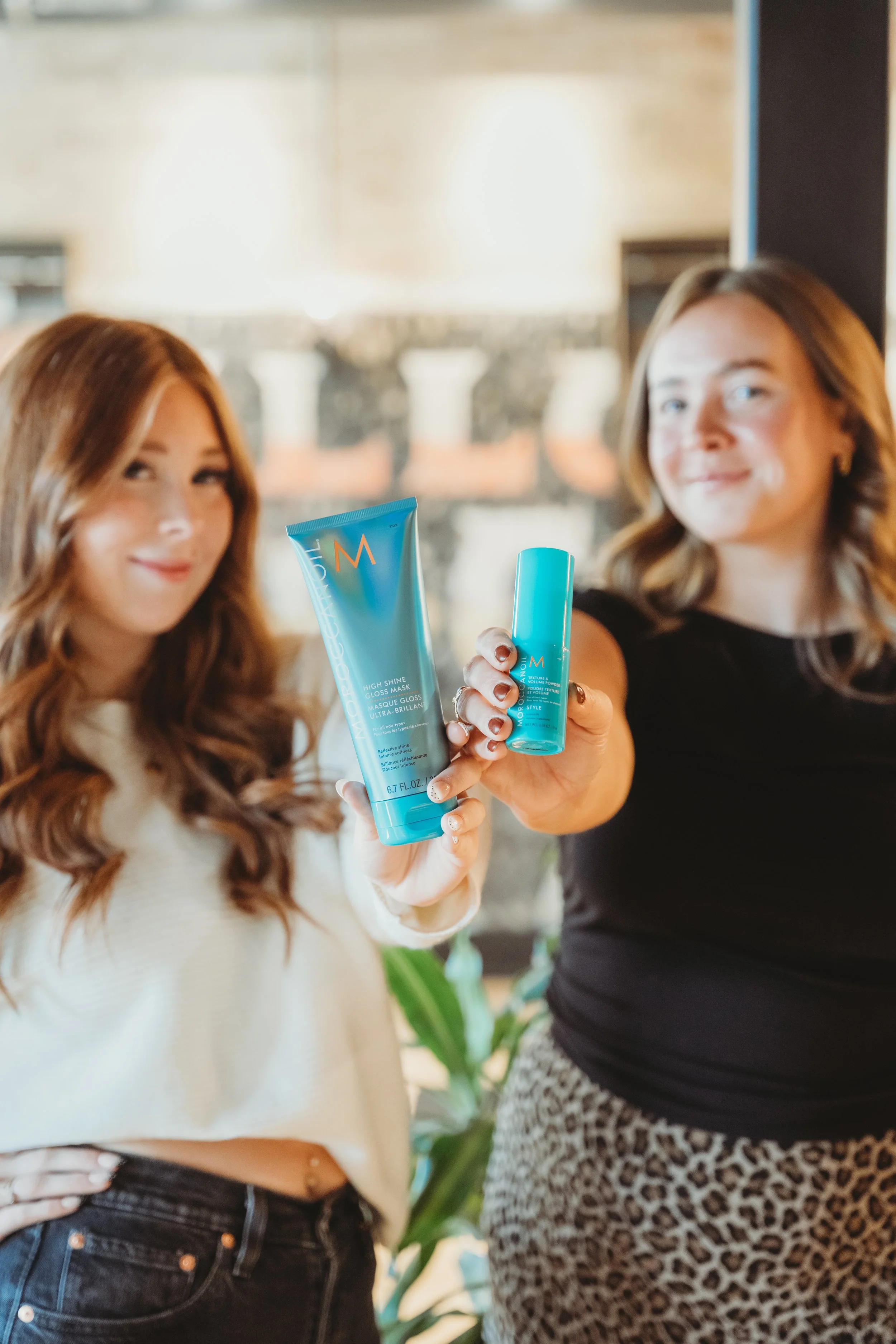 Two women holding blue skincare products, smiling at the camera, inside a bright room with a blurred background.