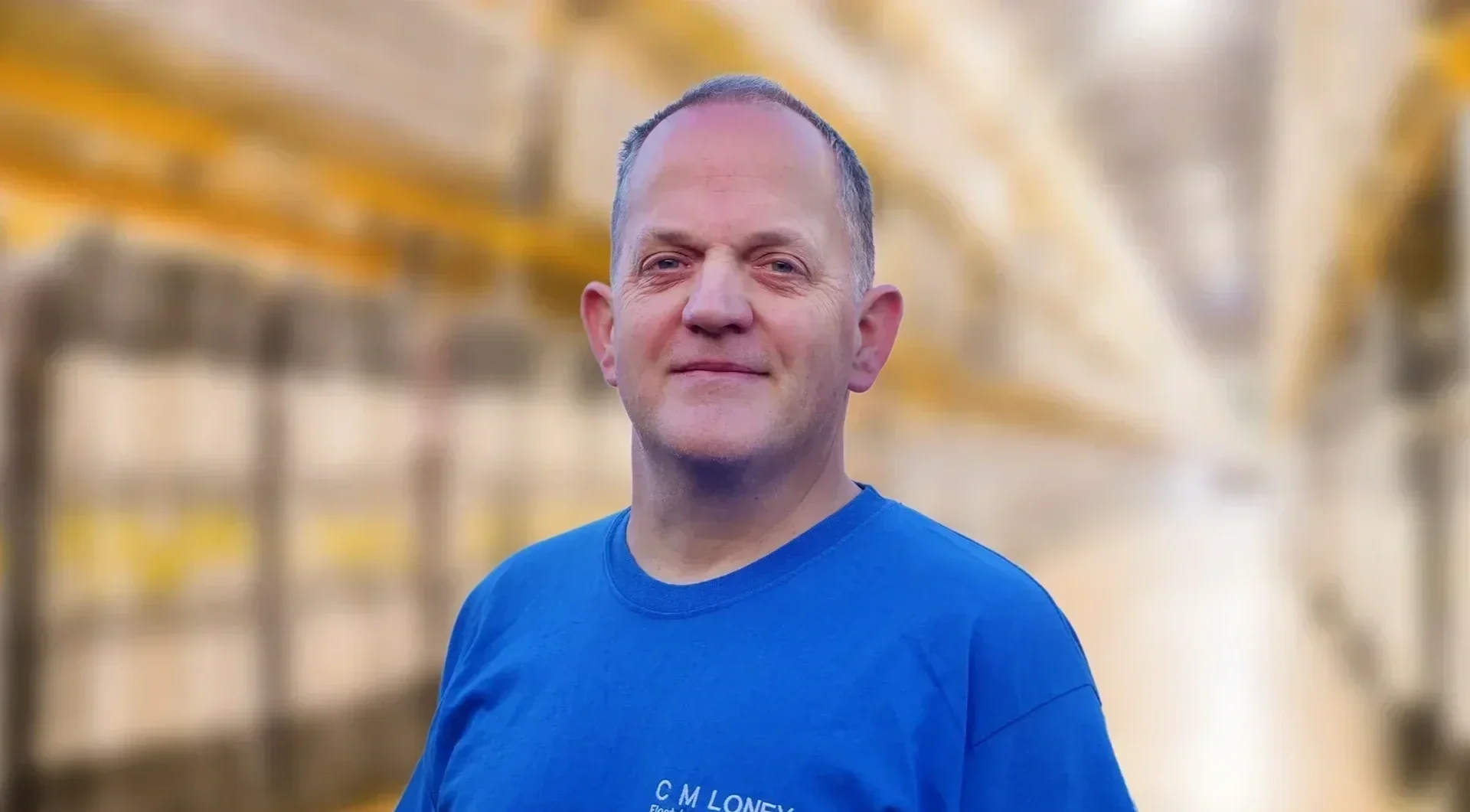 A headshot of owner Chris Loney. Wearing a blue T-shirt with Chris Loney Electrical Services on. Standing in front of a blurred industrial background.